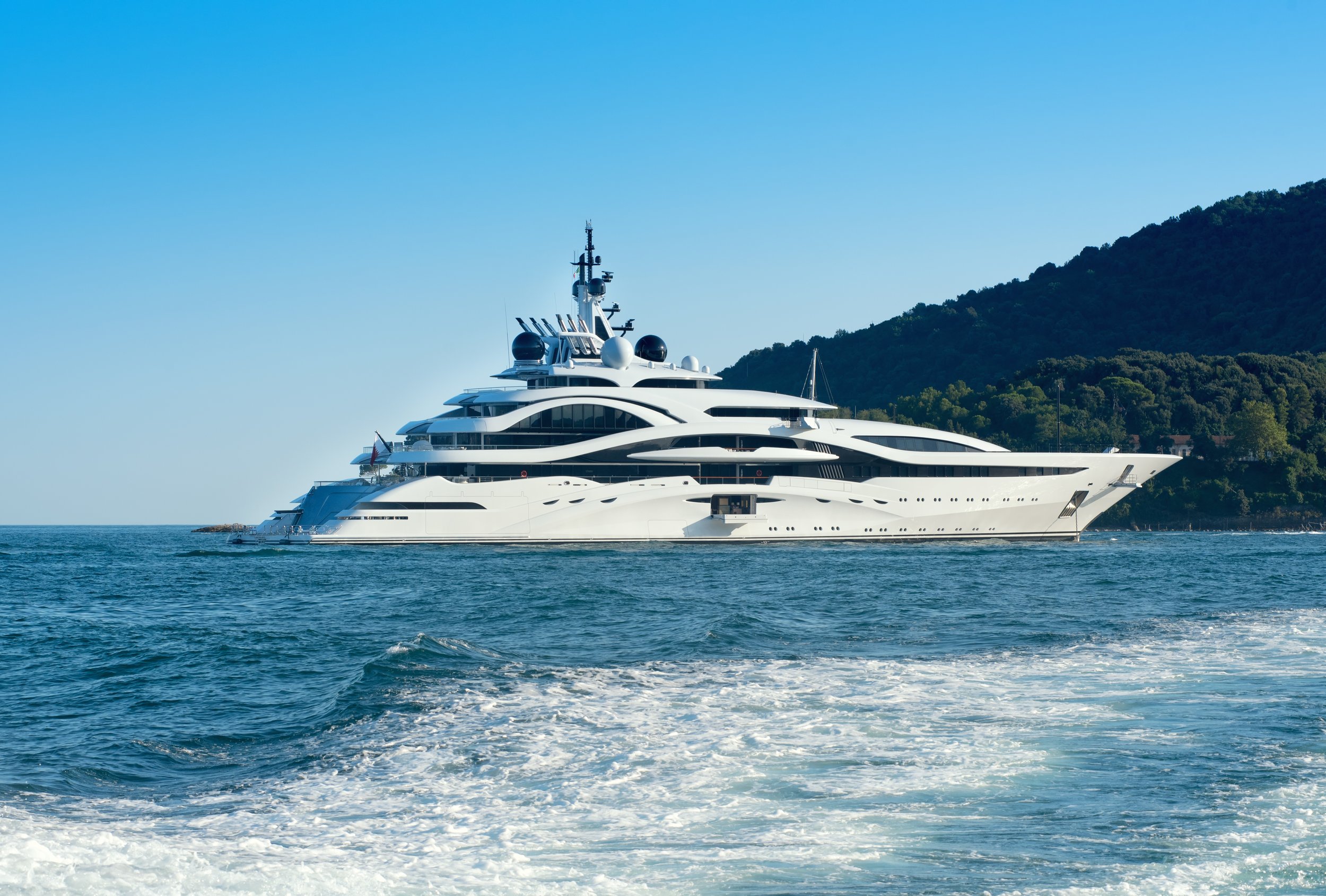 Large white luxury yacht sailing near a green, forested hillside on a clear day.