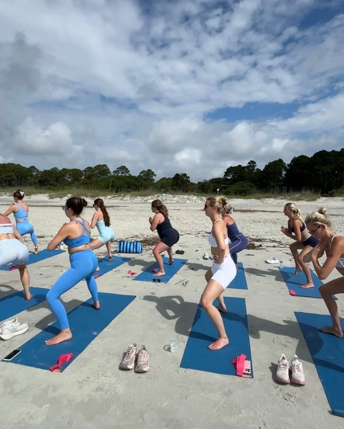 Women practicing yoga on a sandy beach with blue yoga mats under a partly cloudy sky, surrounded by trees.