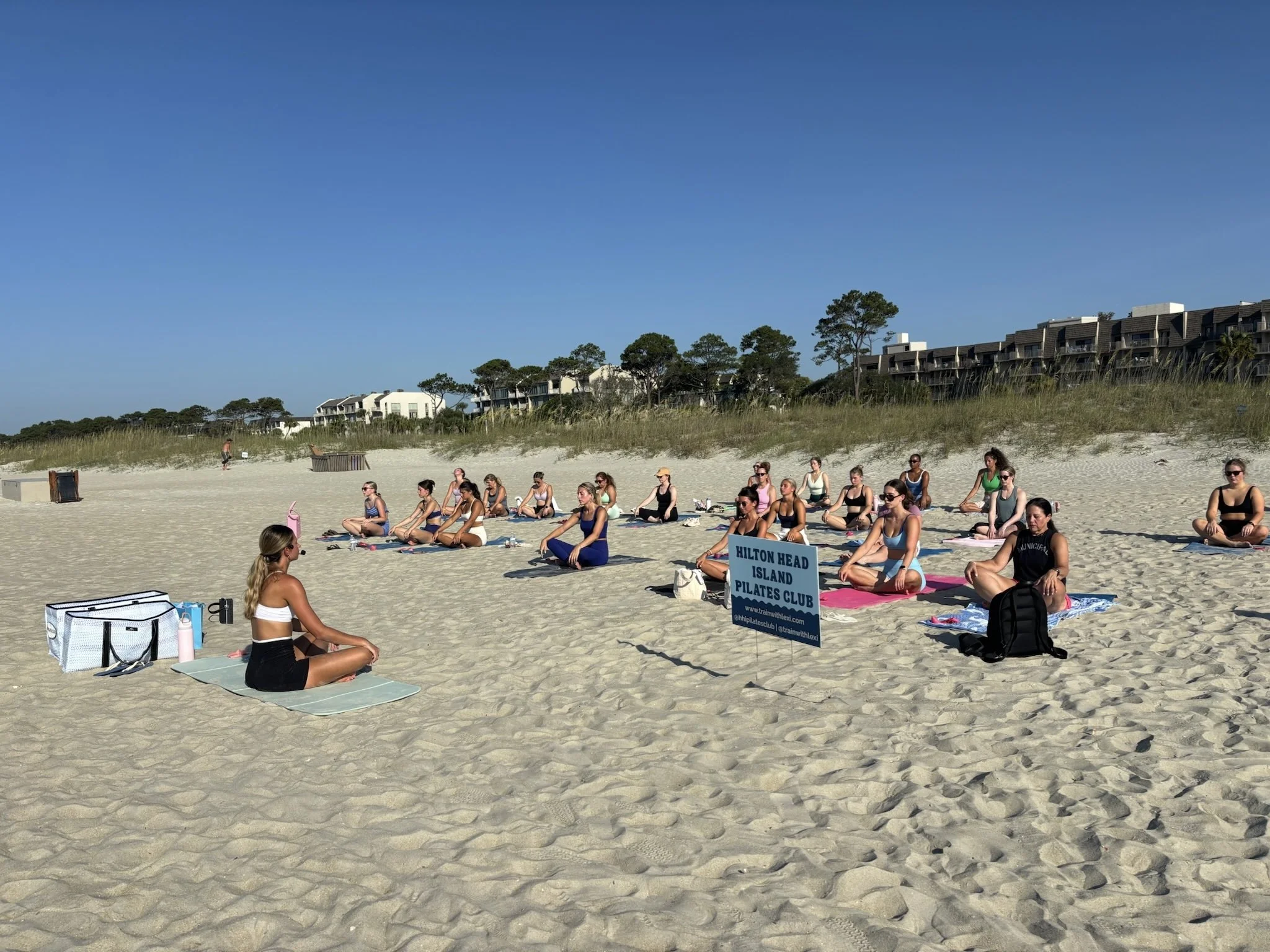 People practicing yoga on the beach during an outdoor yoga class with a sign that reads 'Hilton Head Island Pilates Club' in the foreground.