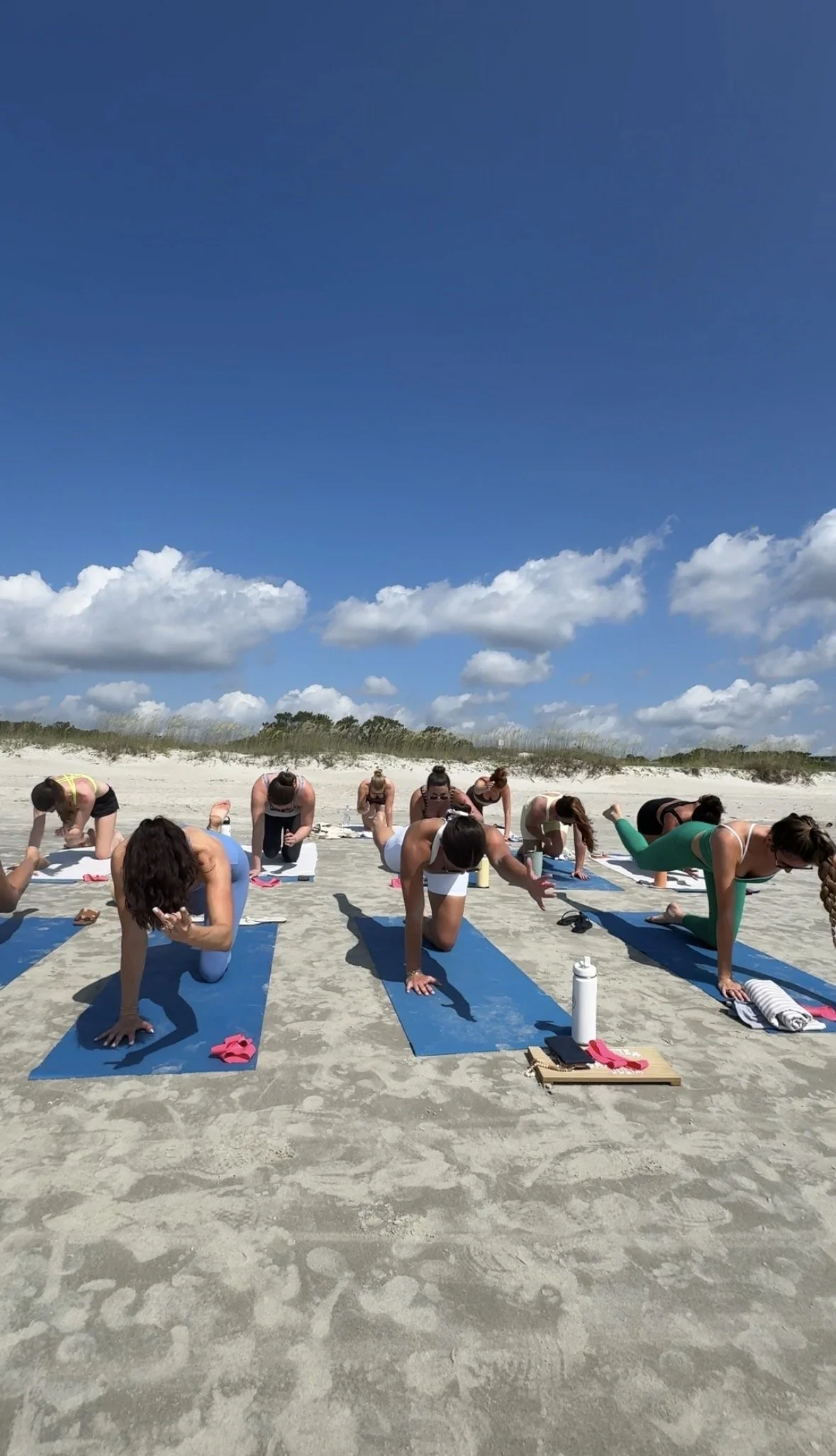 Group of women practicing Pilates on blue mats on the sand at the beach under partly cloudy sky