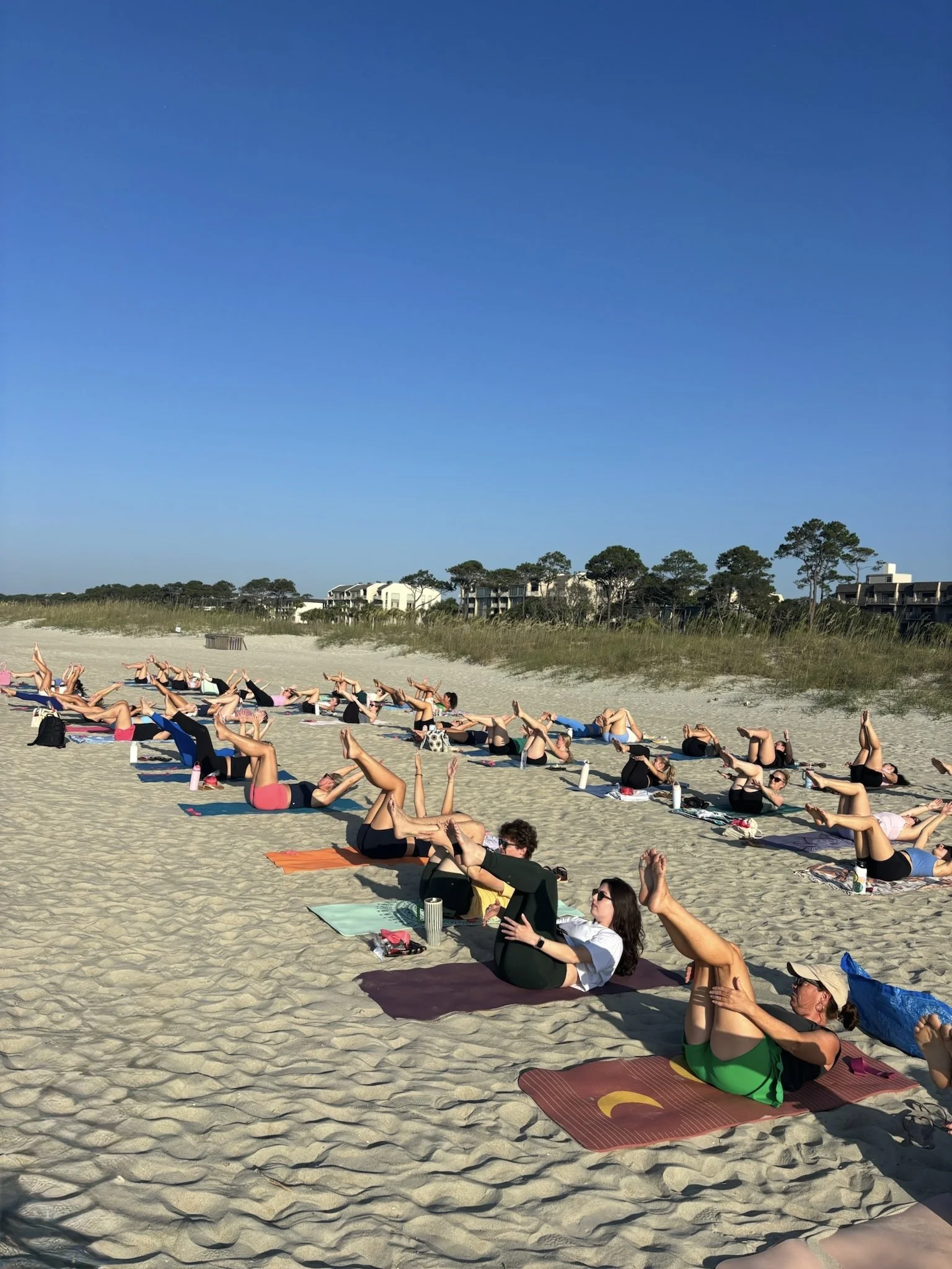 People practicing yoga on a beach with blue sky, sand, and houses in the background.