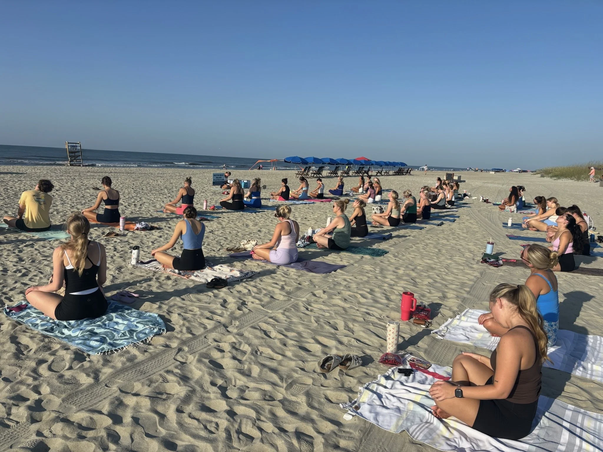 People practicing yoga on the beach during sunrise or sunset, sitting on yoga mats in a meditative pose facing the ocean, with umbrellas and people in the background.