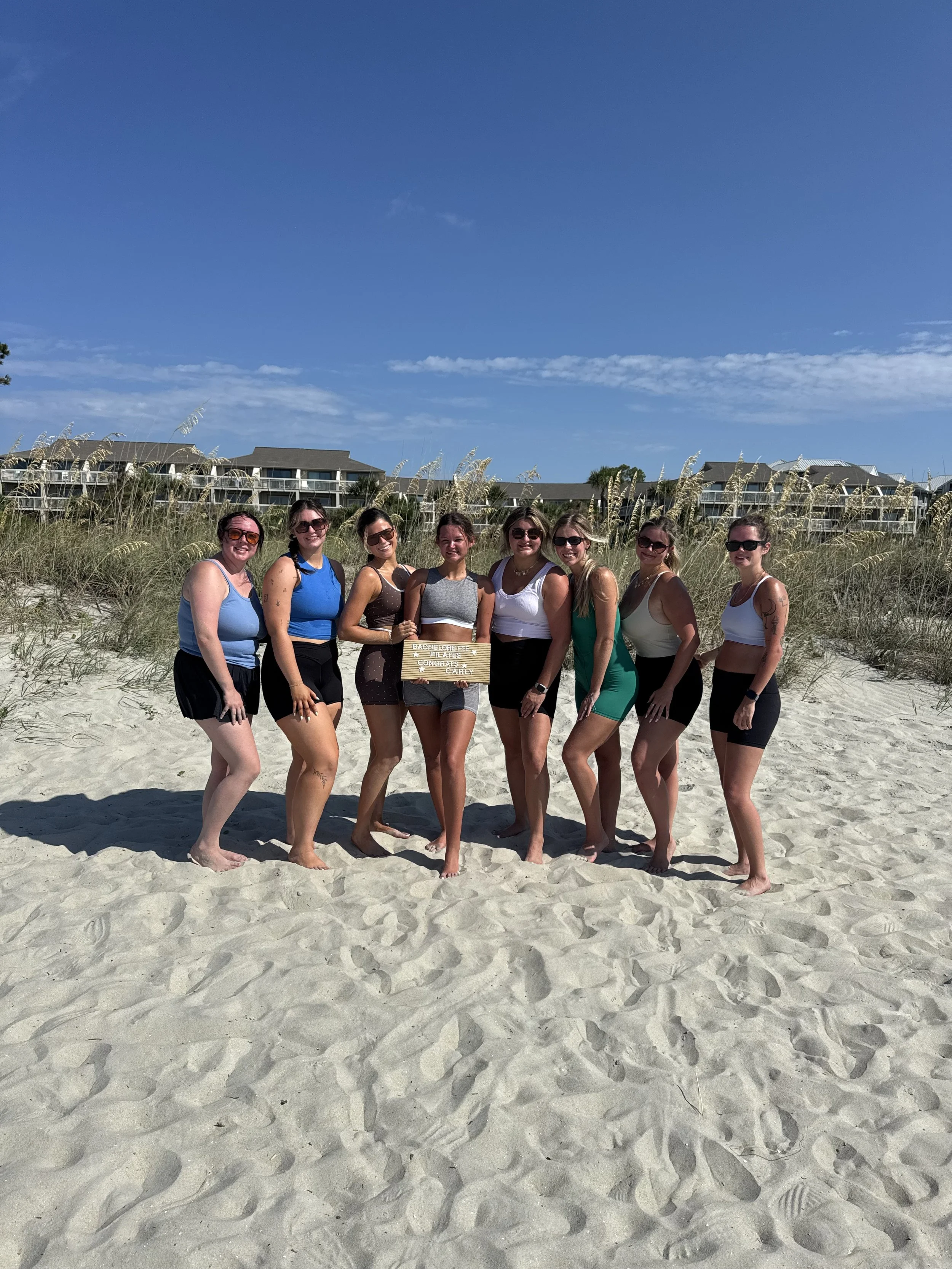 Group of nine women standing on sandy beach with grass and buildings in background, holding a small sign that says "Bachelorette Team" and smiling at the camera.