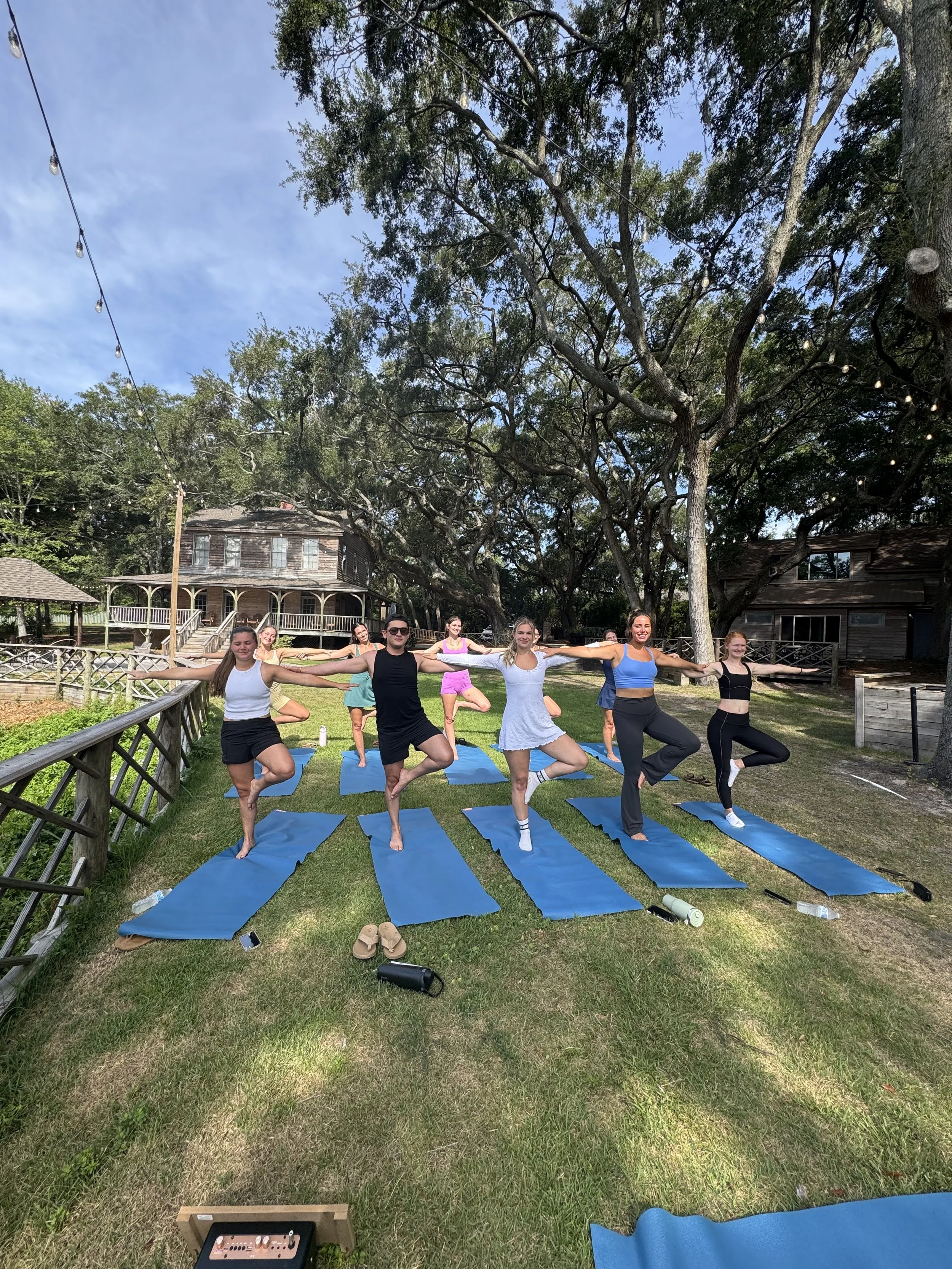 A group of people practicing yoga on blue mats outdoors in a grassy area under large trees with strings of lights hanging above. In the background, there are rustic wooden houses.