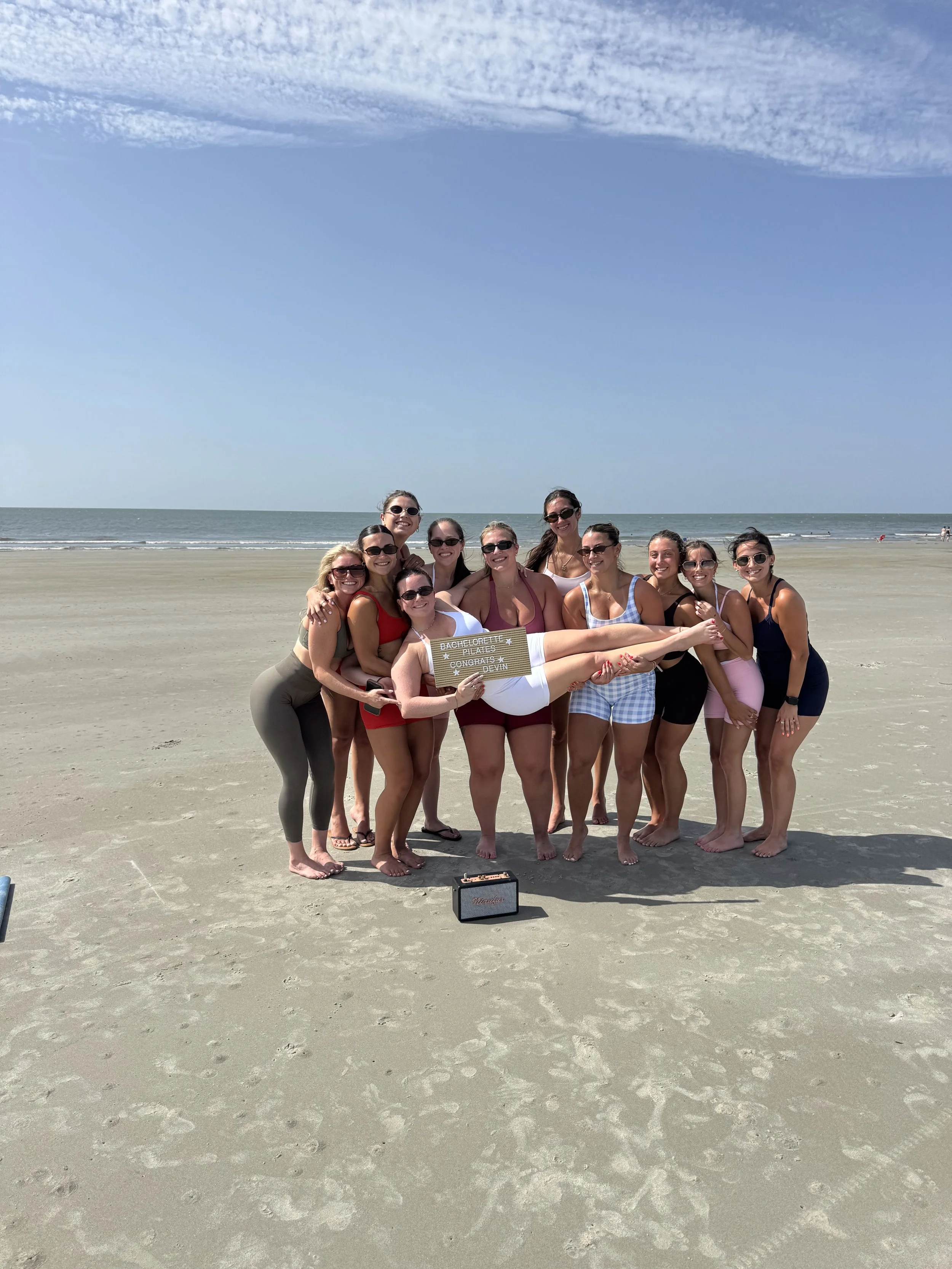 A group of eleven women on a beach, some wearing sunglasses and colorful clothing, holding a sign that reads "Bachelorette Pilates" and "Congrats Devin". One woman is lying across the others while holding a vintage speaker on the sand. The sky is clear with some clouds, and the ocean is in the background.