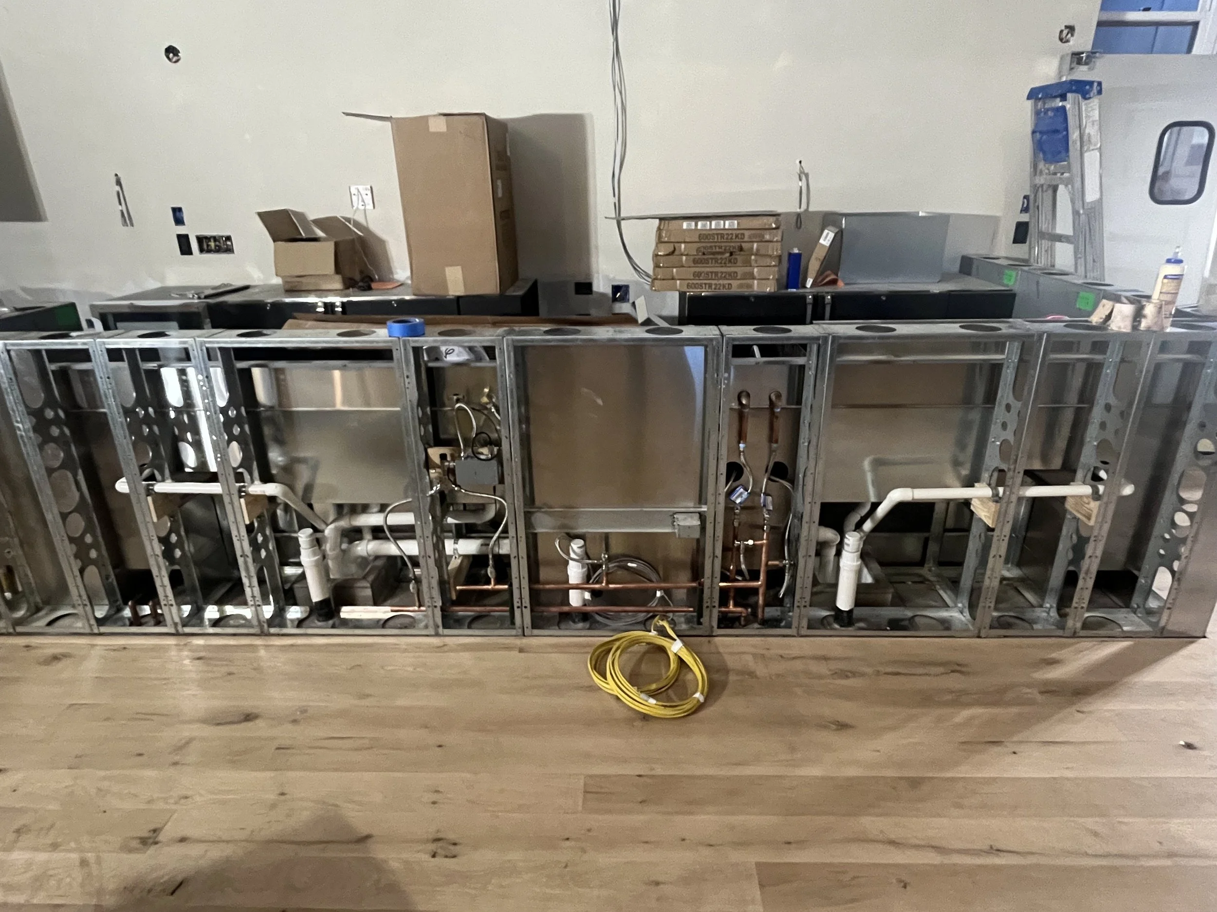 Unfinished commercial kitchen appliance installation with metal framework, copper and PVC piping, and cardboard boxes and ladders in the background.