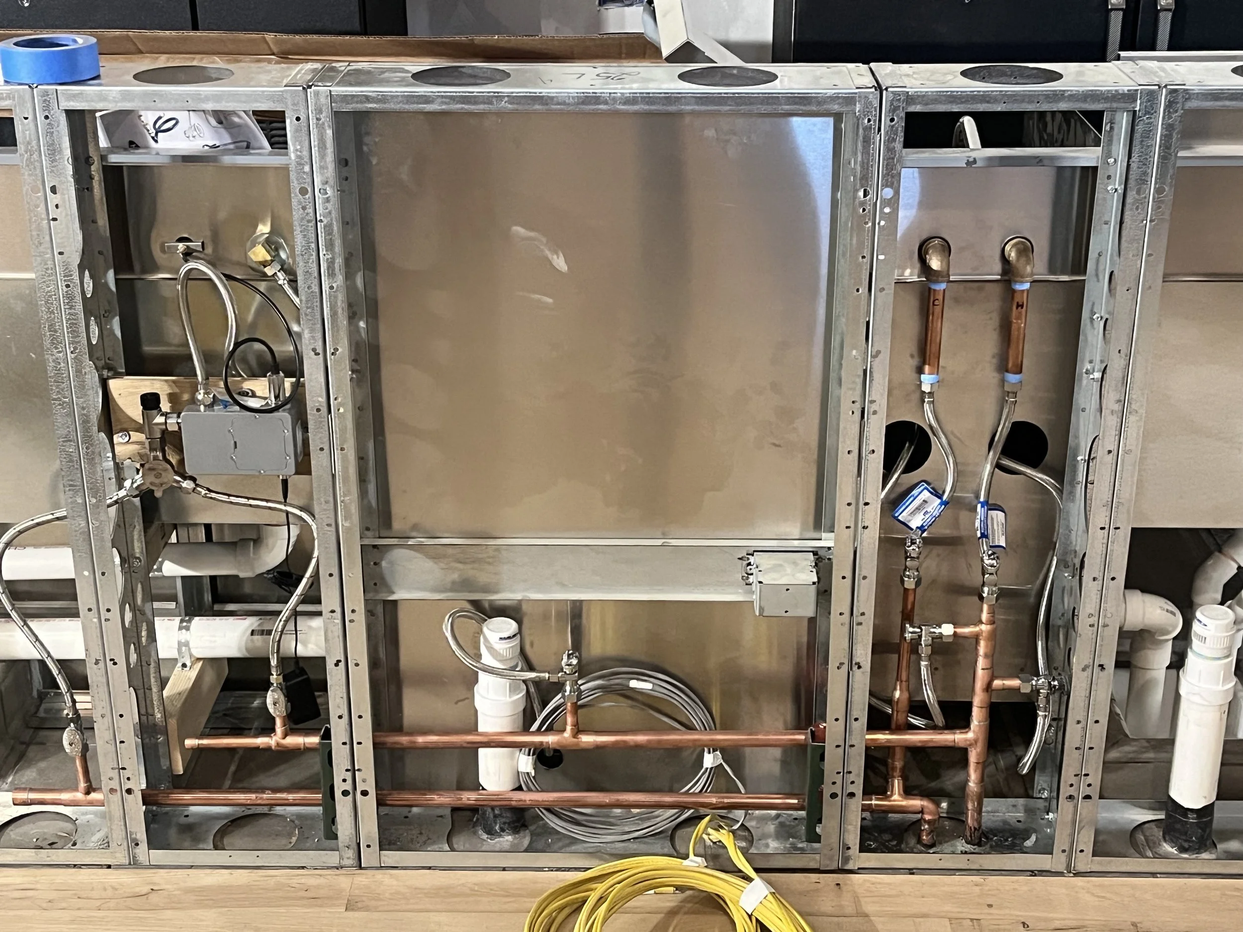 Metal framing with plumbing components, copper pipes, PVC pipes, and electrical wiring for a kitchen or bathroom fixture installation.
