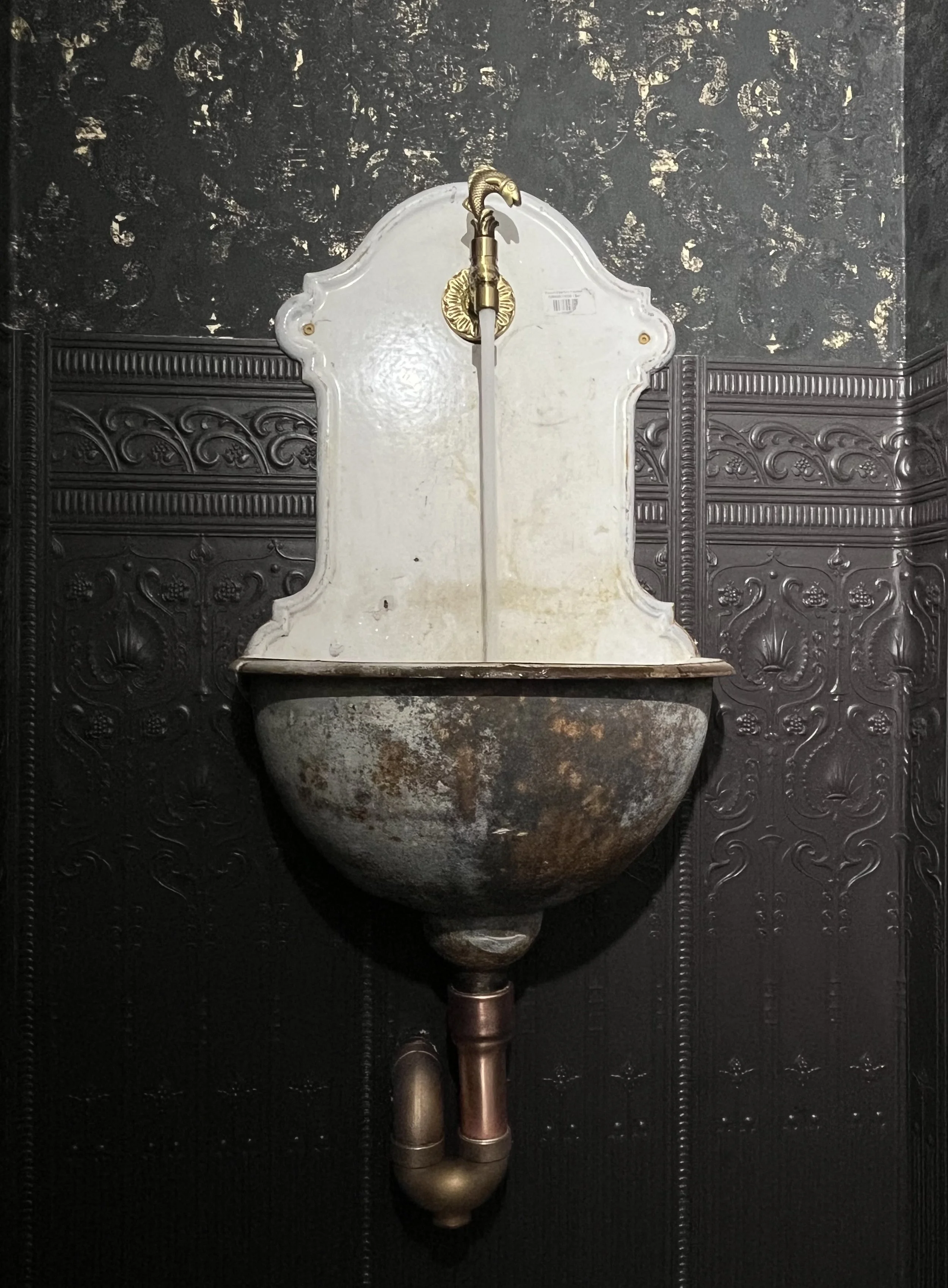Vintage wall-mounted sink with a rusty basin, ornate white backplate, and a brass faucet shaped like a swan, set against decorative black embossed wall panels.