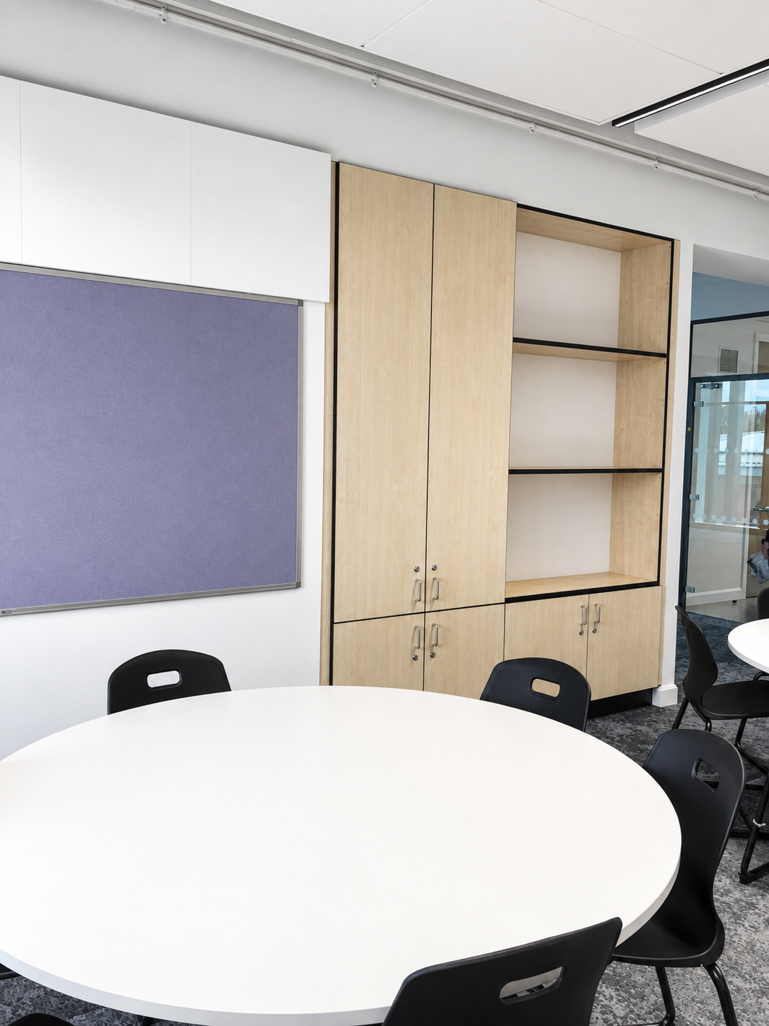 Classroom at Peebles High School featuring bespoke fitted timber storage cabinets, open shelving, a wall-mounted pinboard and circular tables.
