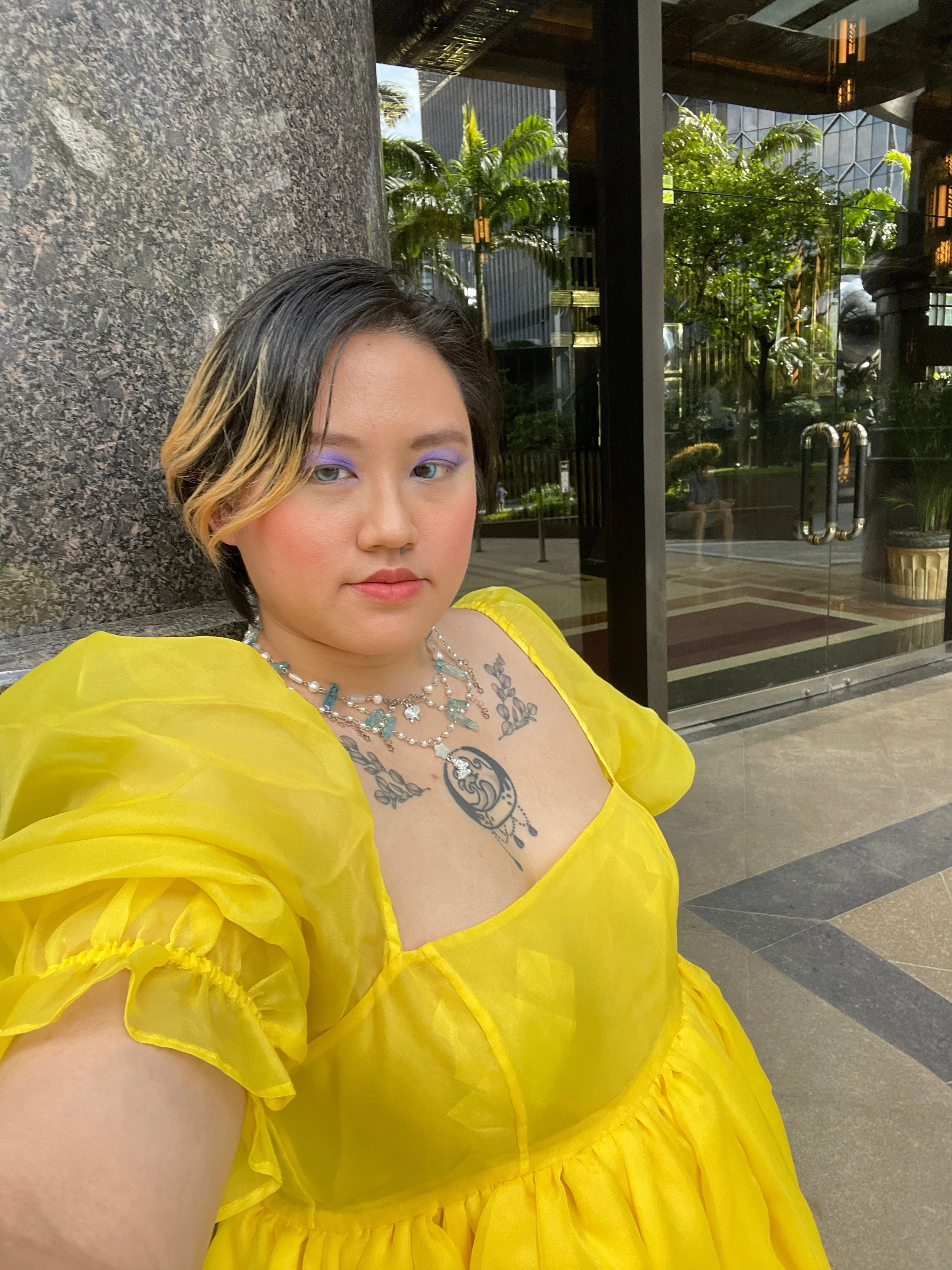 Person wearing a yellow dress with puffed sleeves, purple eyeshadow, and a necklace, standing near a structure with greenery and glass doors in the background.
