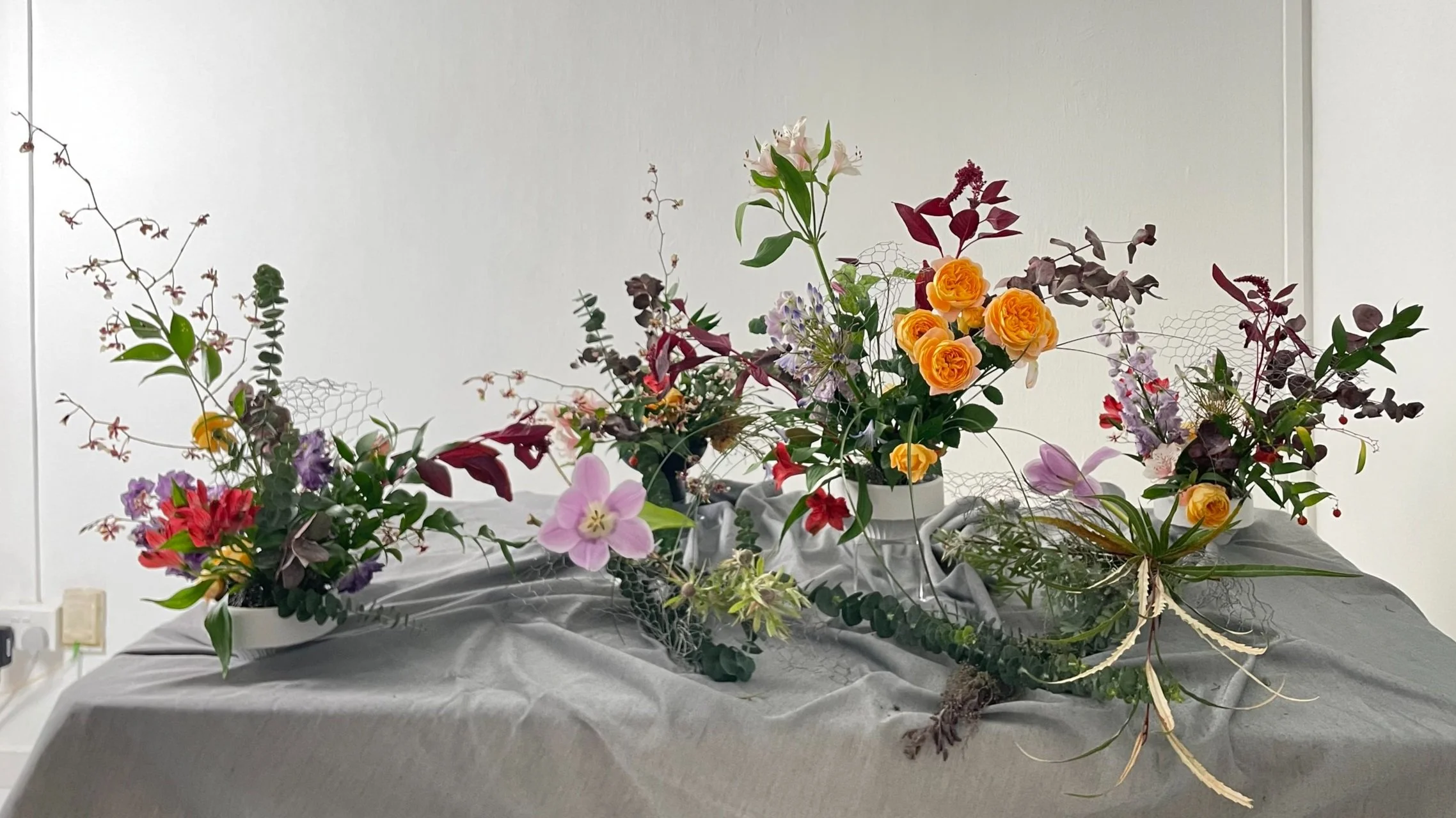 Arrangements of flowers including roses, lilies, and various green and purple foliage, displayed on a draped gray fabric.