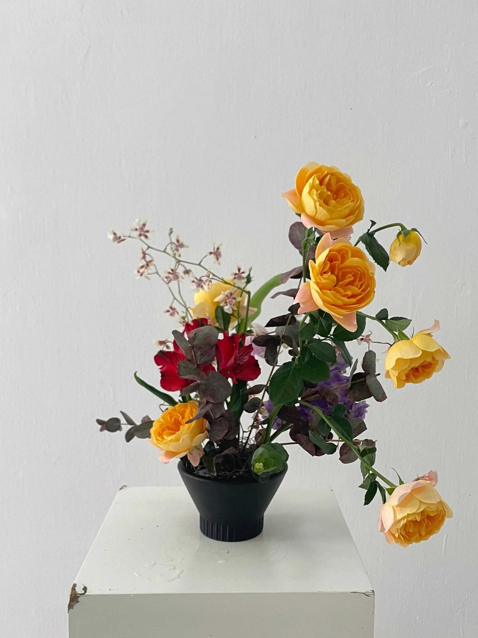 Ikebana floral arrangement with yellow roses and red flowers in a black vase on a white pedestal.