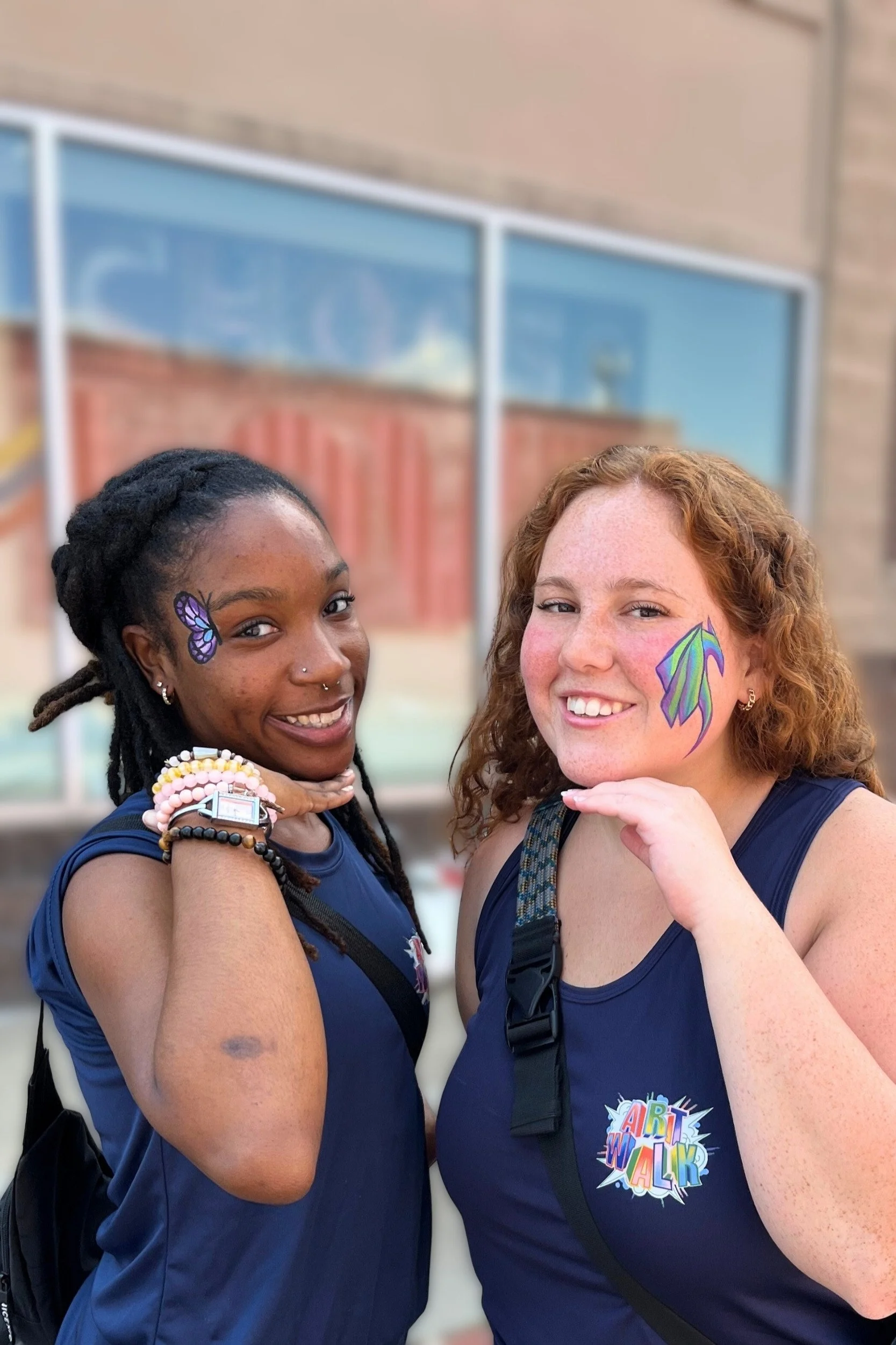 Two staff members standing outdoors, smiling, with face paint, wearing matching navy blue shirts, one with a butterfly face painting and the other with a colorful feather design on her cheek.
