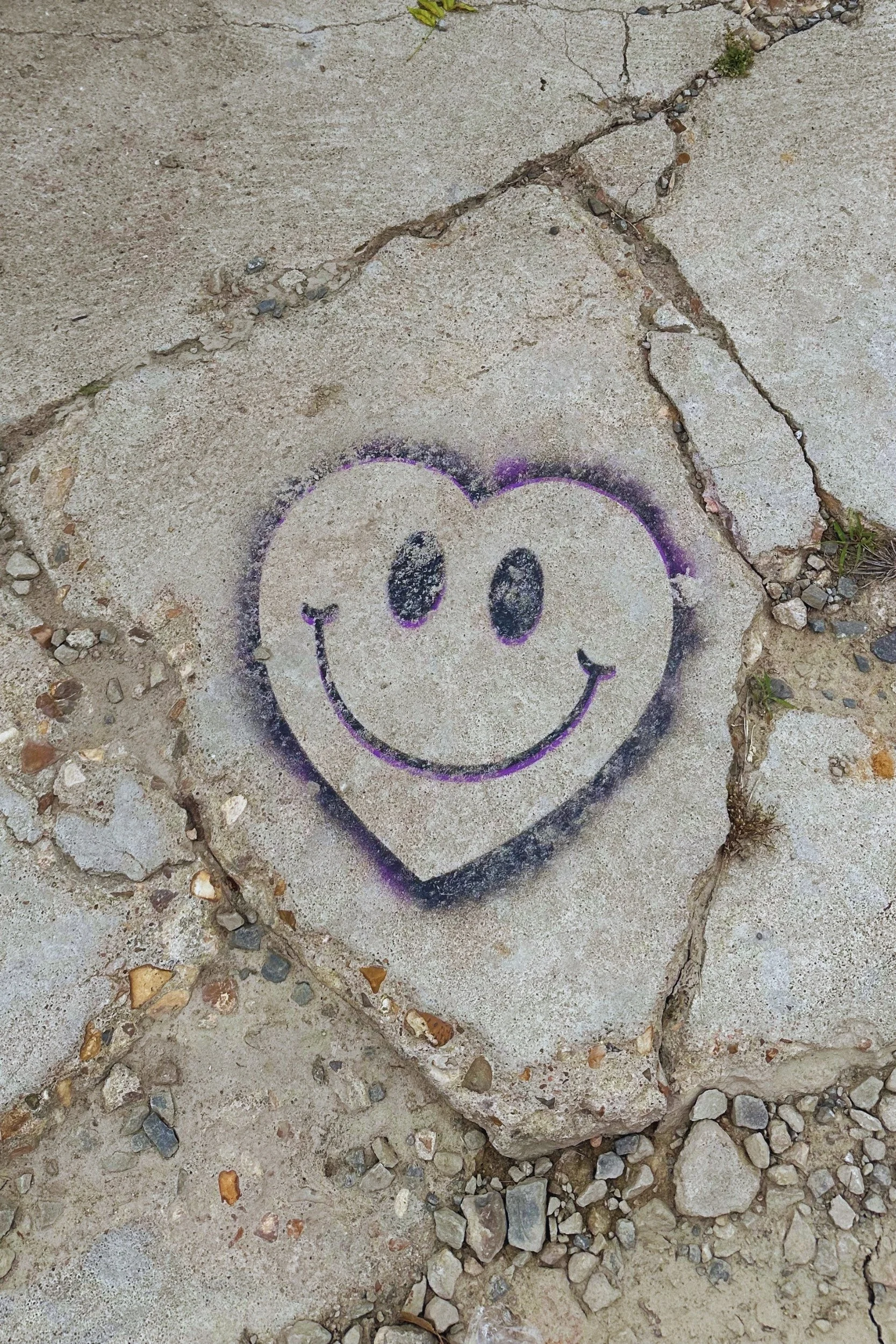A smiley face drawn in a heart shape with a black and purple outline over cracked concrete