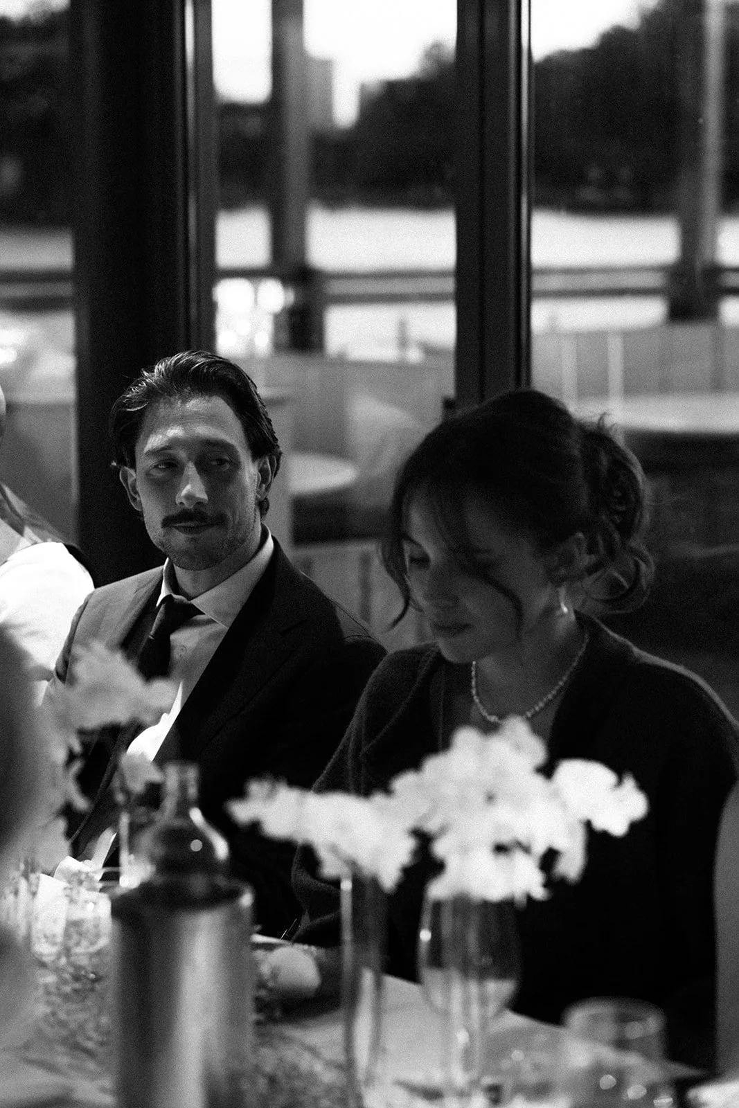 Een zwart-wit foto van een man en vrouw die aan een tafel zitten. De man draagt een pak en ziet er ontspannen uit, terwijl de vrouw naar beneden kijkt en een ketting draagt. Op de tafel staan een bloemenvaas en een flesje.