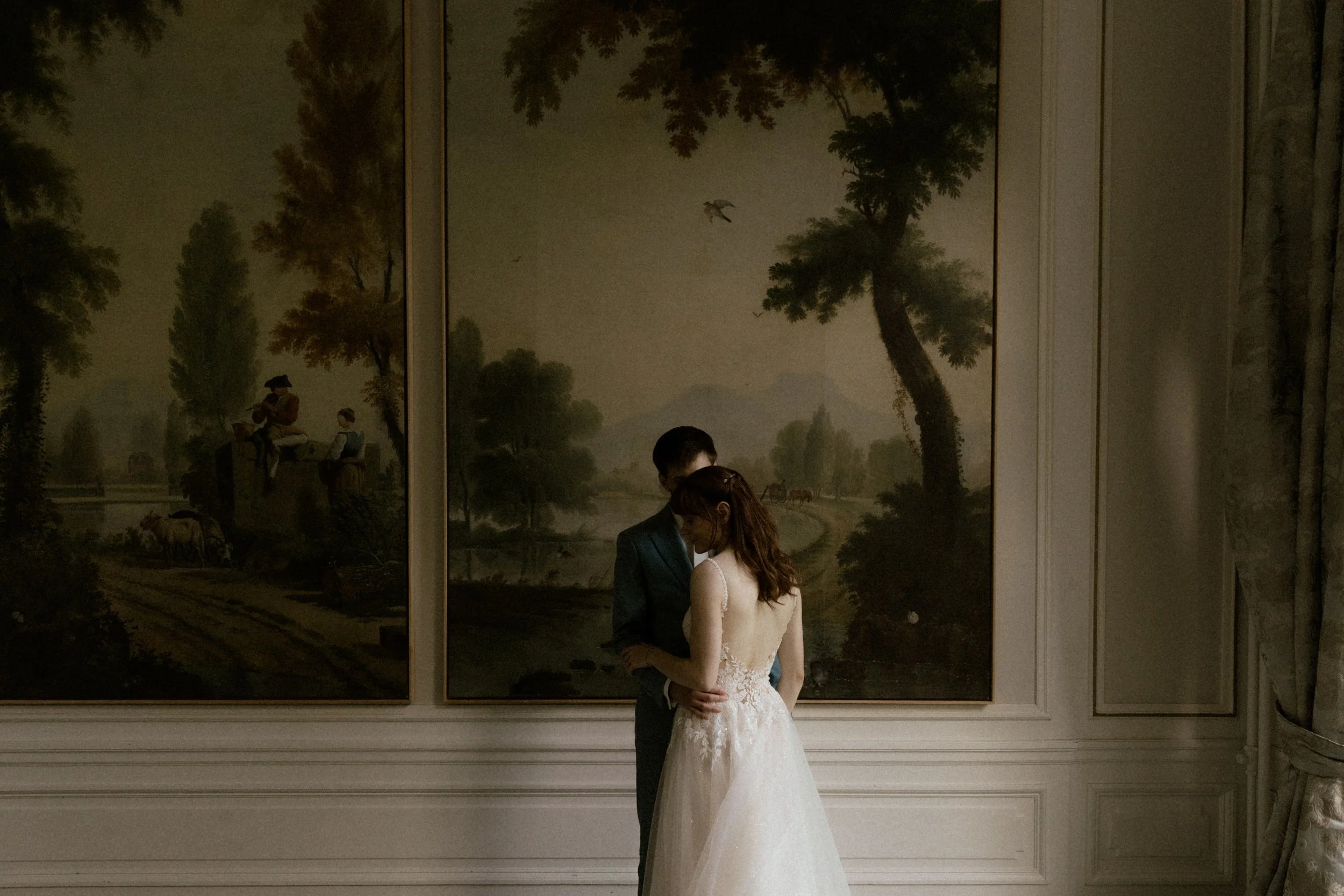 Een koppel in bruidskleding staat dicht tegen elkaar aan in een elegant interieur met wandkunst, mogelijk tijdens hun bruiloft.