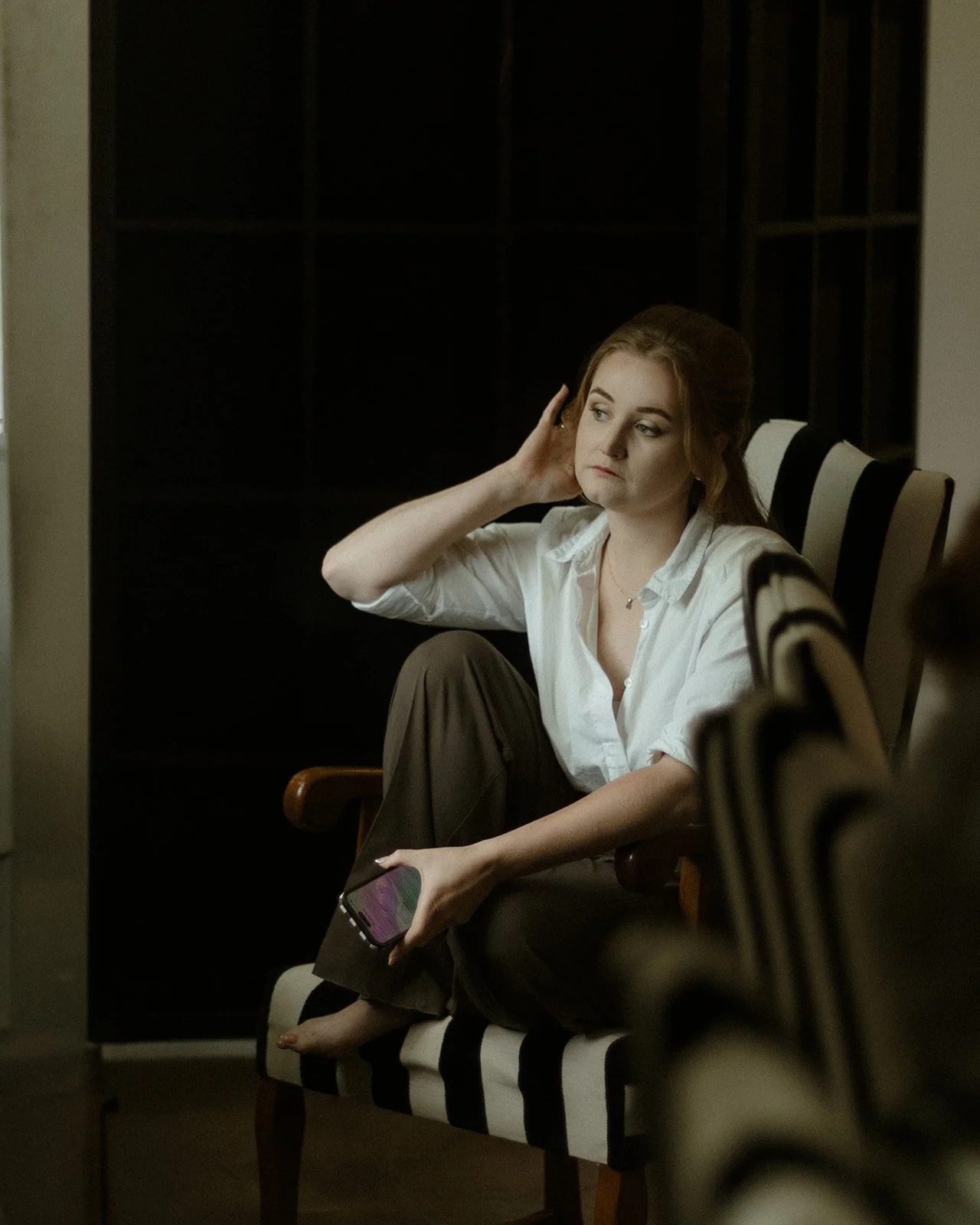 Een jonge vrouw zit op een bank met zwarte en witte strepen, in een donkere kamer, terwijl ze haar hoofd aan haar hand ondersteunt en naar haar telefoon kijkt, met een serieuze uitdrukking.