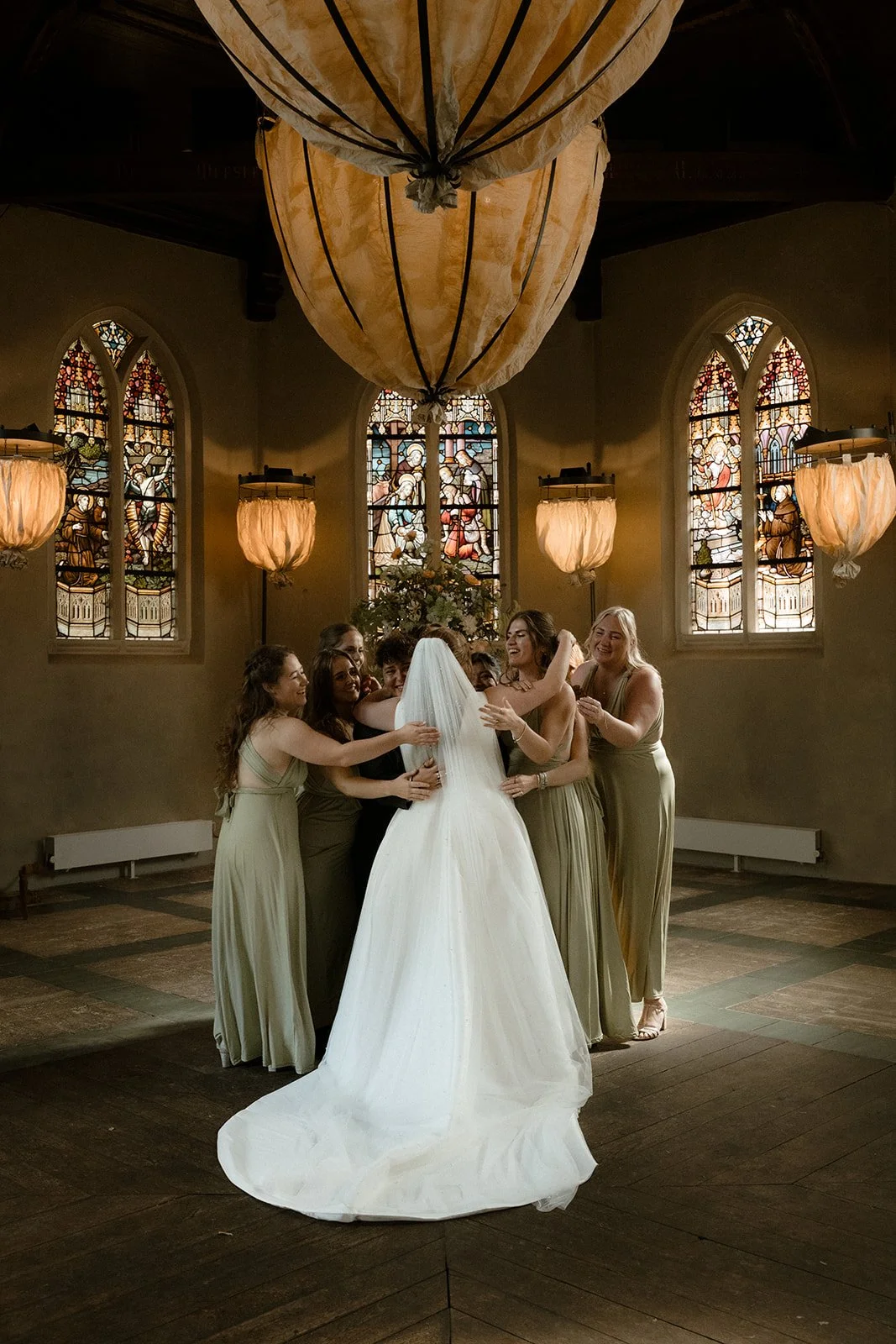 Een groep vrouwen die elkaar omhelzen en lachen tijdens een huwelijk in een kerk met stained glass ramen.