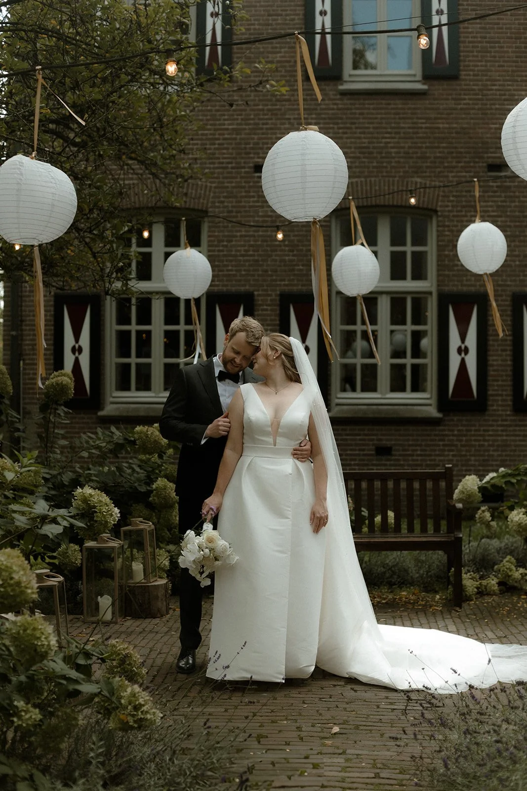 Een getrouwd stel in een tuin met papieren lantaarns boven hen, omringd door groene planten en bloemen, voor een bakstenen gebouw met ramen en decoratieve luiken.