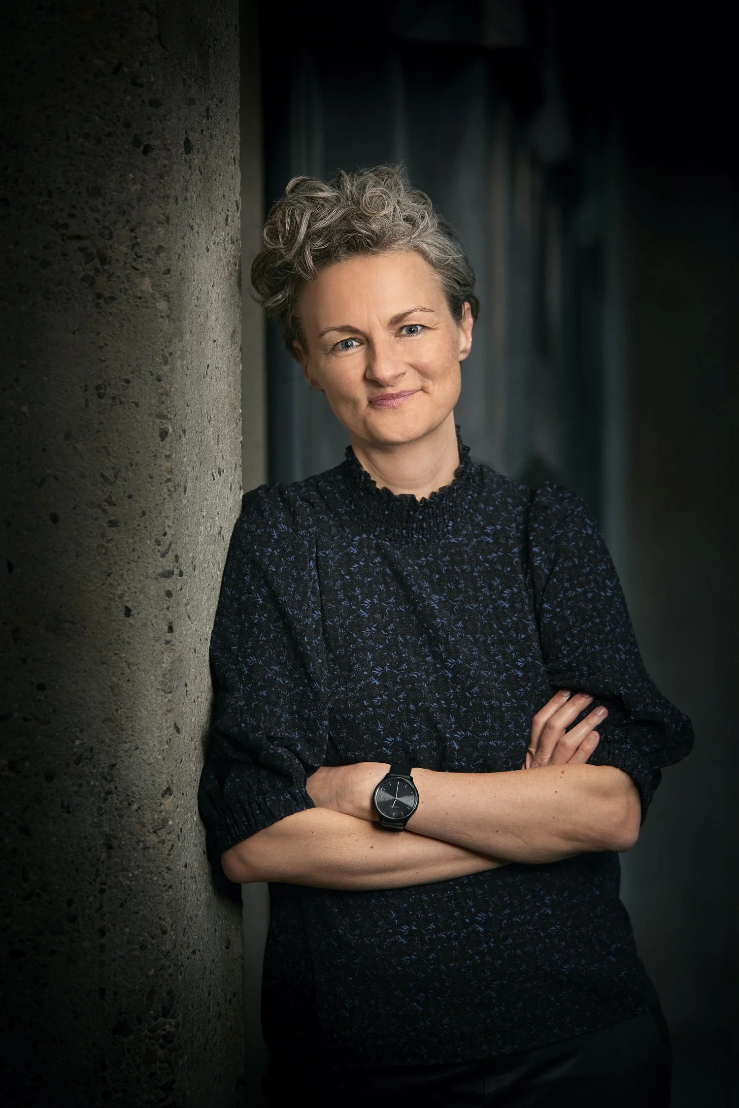 Laura Vilsbæk med gråt, krøllet hår, iført en mørk, langærmet bluse med en lille blomsterprint, der står med armene krydsede ved en væg.