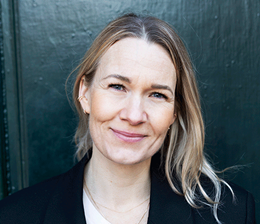 Kvinde med blond, bølget hår, iført en grøn polo med logo, smilende, mod baggrund af grønne blade.