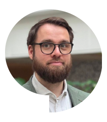 Mand med brunt hår, skæg og briller iført en grå blazer og hvid skjorte, stående indendørs.