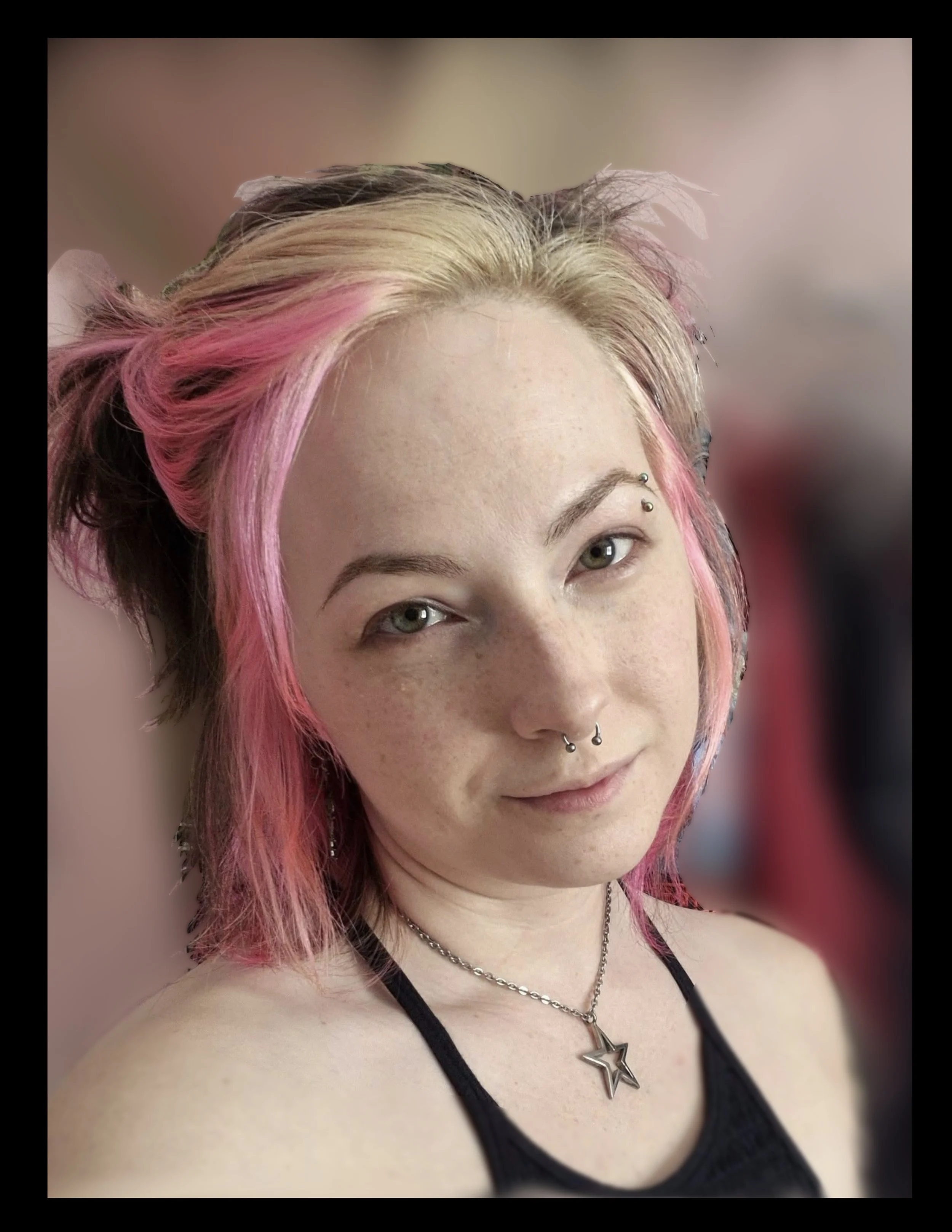 Young woman with long wavy hair taking a selfie, winking, with tattoos on her arm and various artwork on the wall behind her including a clock with a cat face design and framed pieces of art.