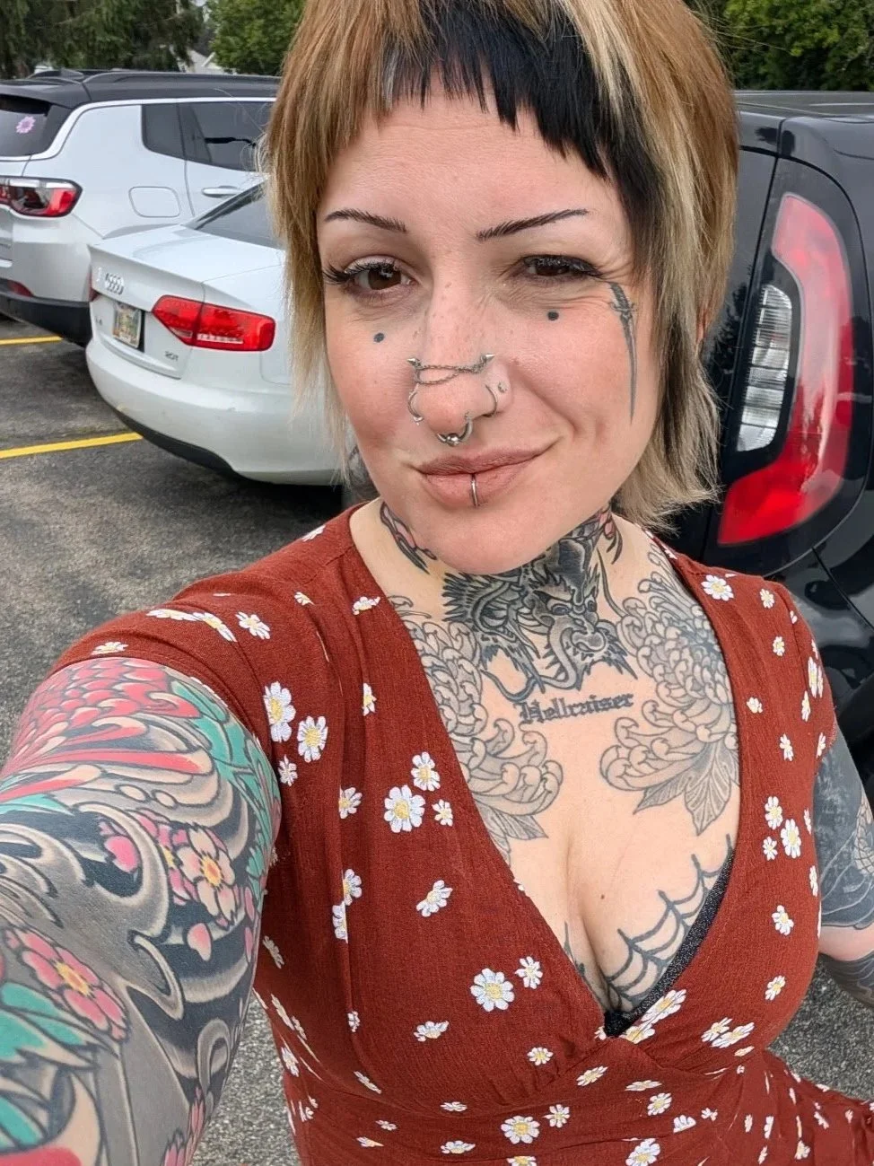 A woman with short hair and numerous tattoos, including flowers and a dragon, taking a selfie in a parking lot with parked cars in the background.