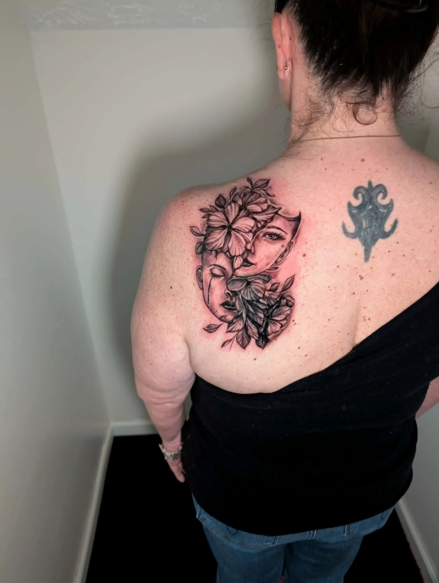 Back of a woman with tattoos on shoulder and neck, wearing a black tank top and blue jeans, facing a wall.