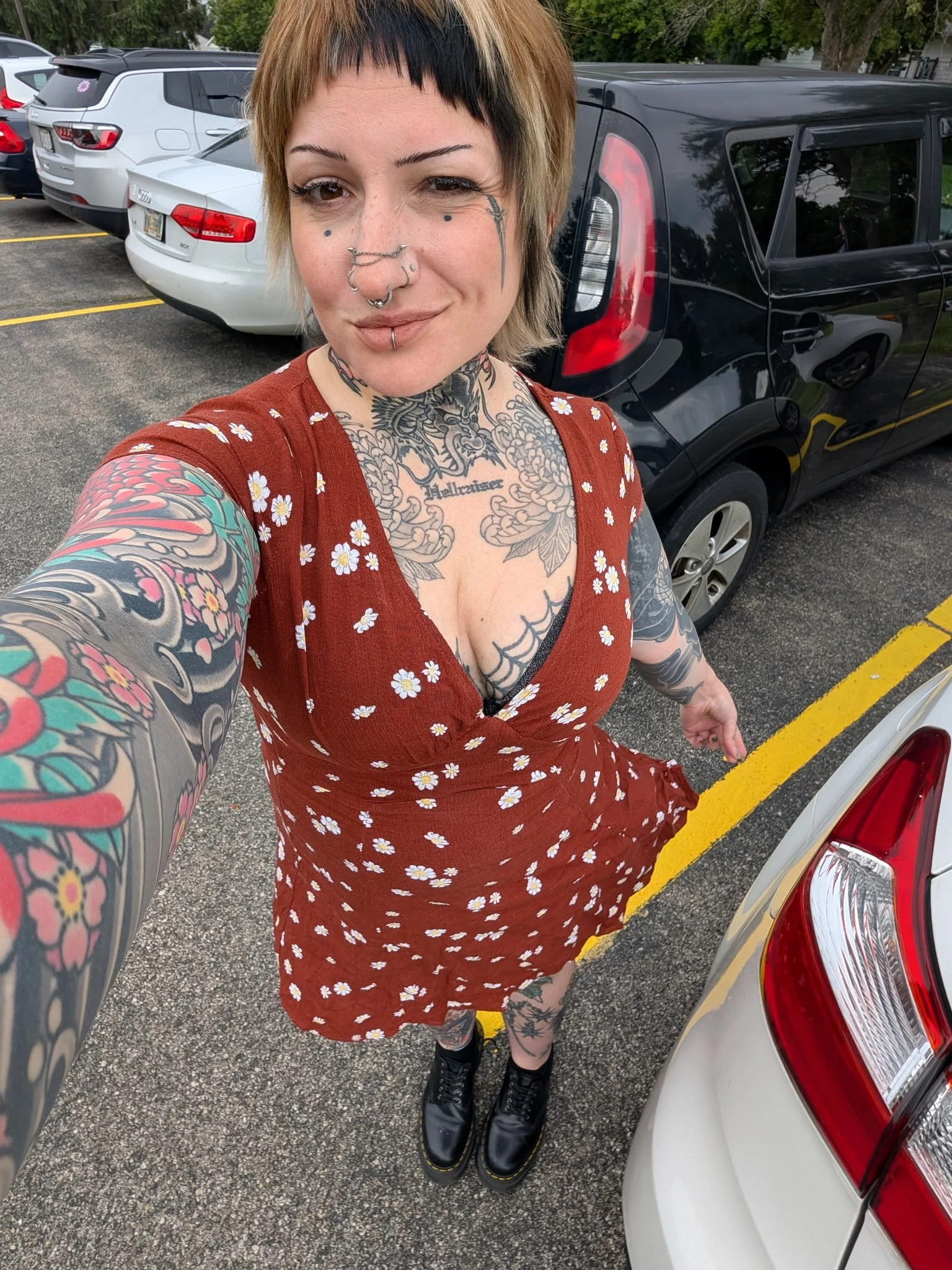 A woman with short hair, multiple tattoos, and facial piercings taking a selfie in a parking lot next to cars, wearing a red dress with a white floral pattern and black boots.