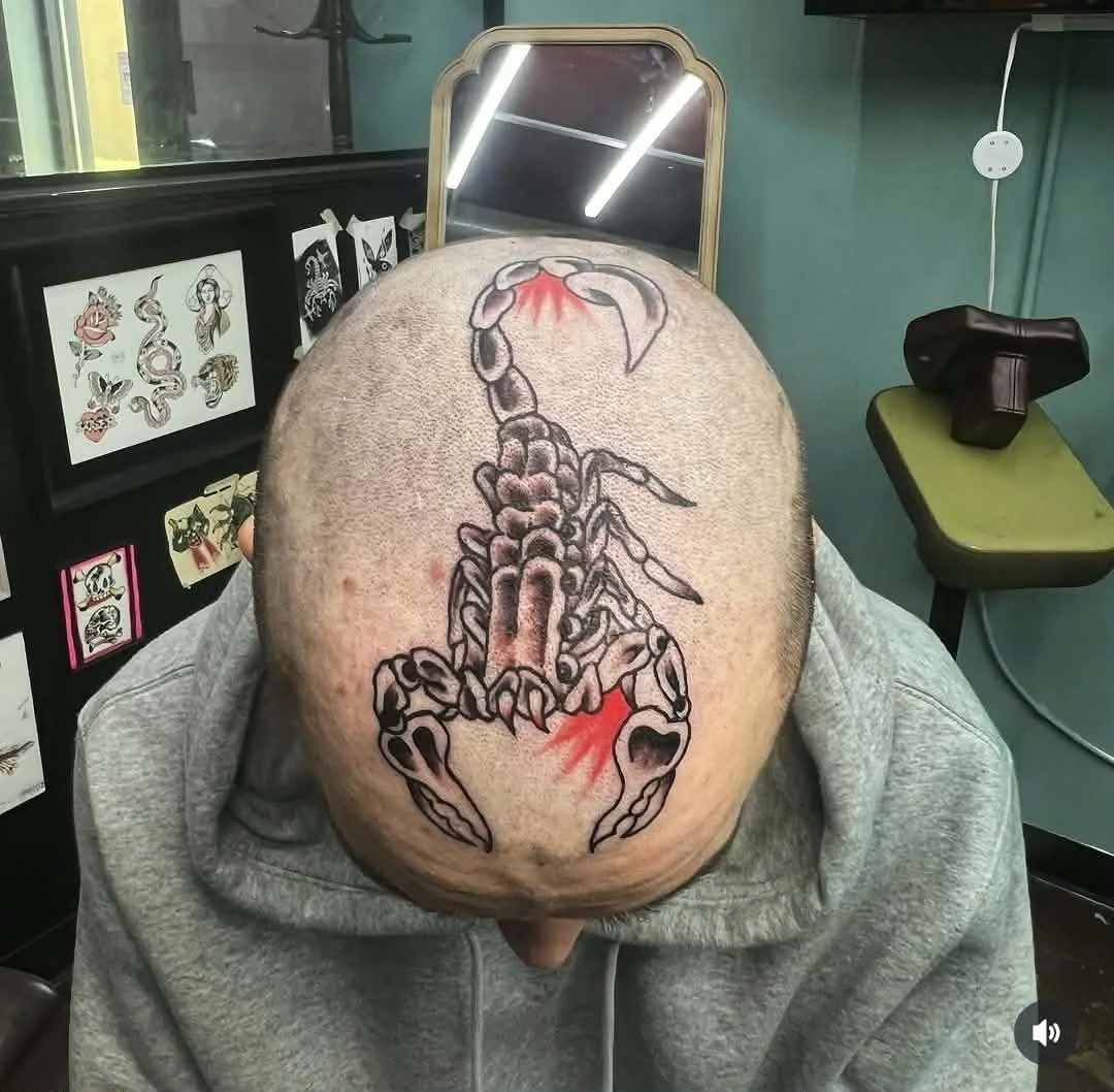 Tattoo of a black and gray scorpion with red accents on a person's bald head, in a tattoo studio with tattoo designs on the wall and a mirror in the background.