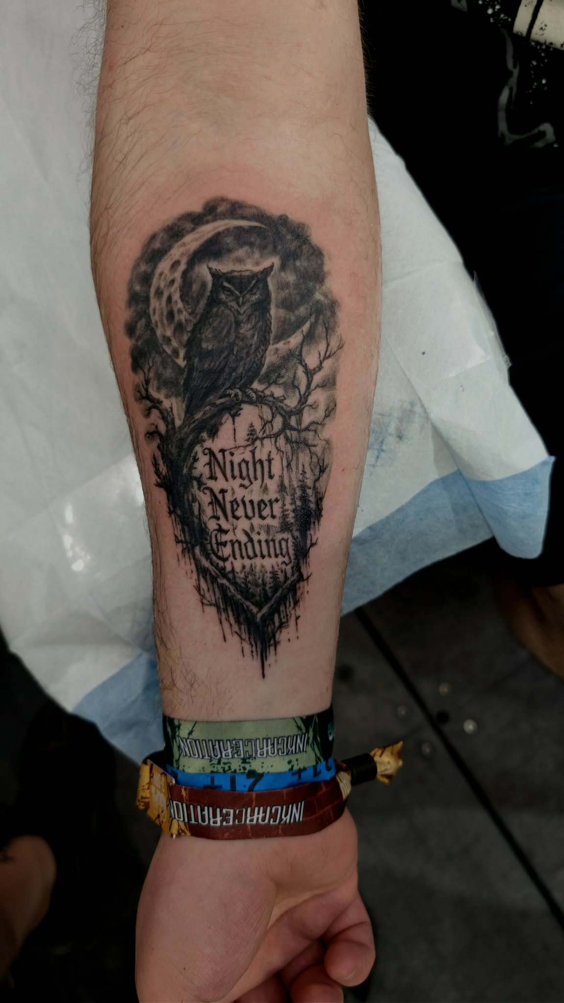 Tattoo of an owl perched on a branch with the phrase "Night Never Ending" written below, featuring a dark, moonlit background with trees.