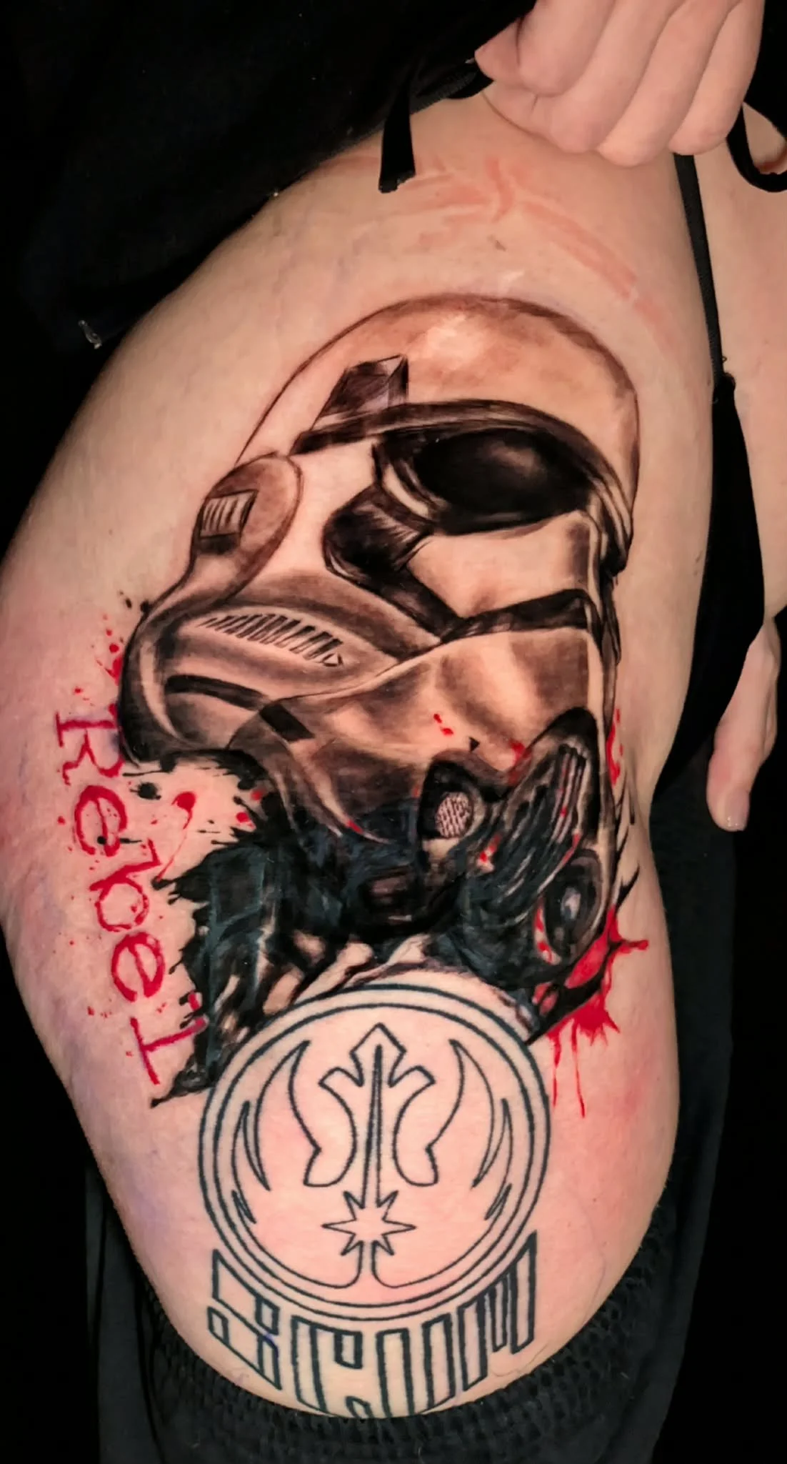 Tattoo of a Stormtrooper helmet from Star Wars on a person's arm, with red and black ink splatters and writing.