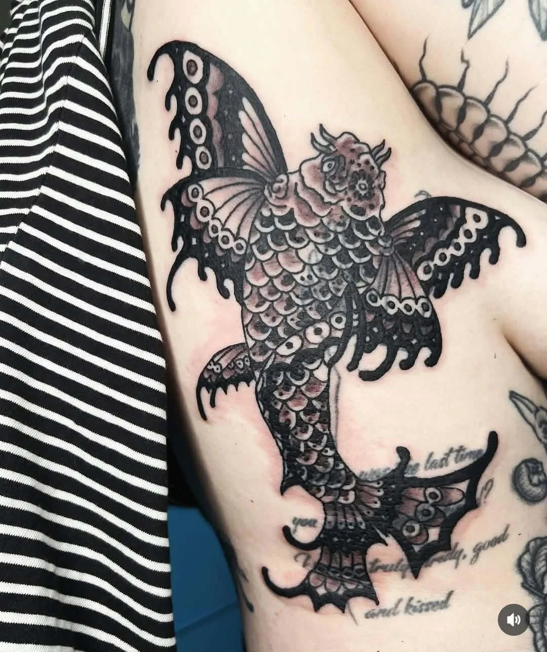 Tattoo of a hybrid creature with butterfly wings and a fish tail on a person's upper arm, with additional text inscriptions.