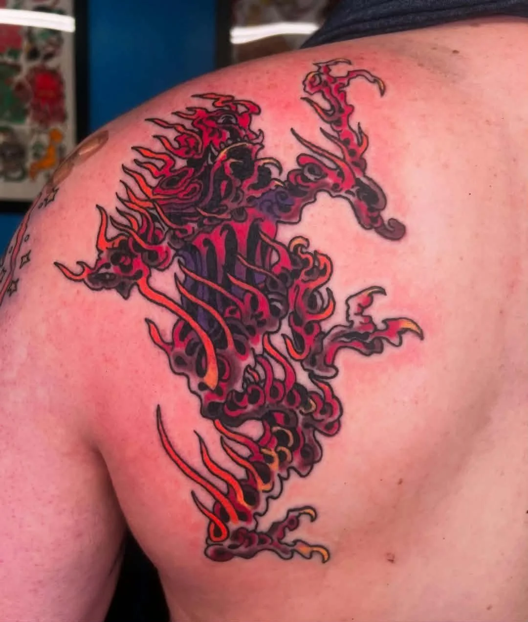 Tattoo of a stylized, fiery dragon in red, black, and purple on a person's shoulder.