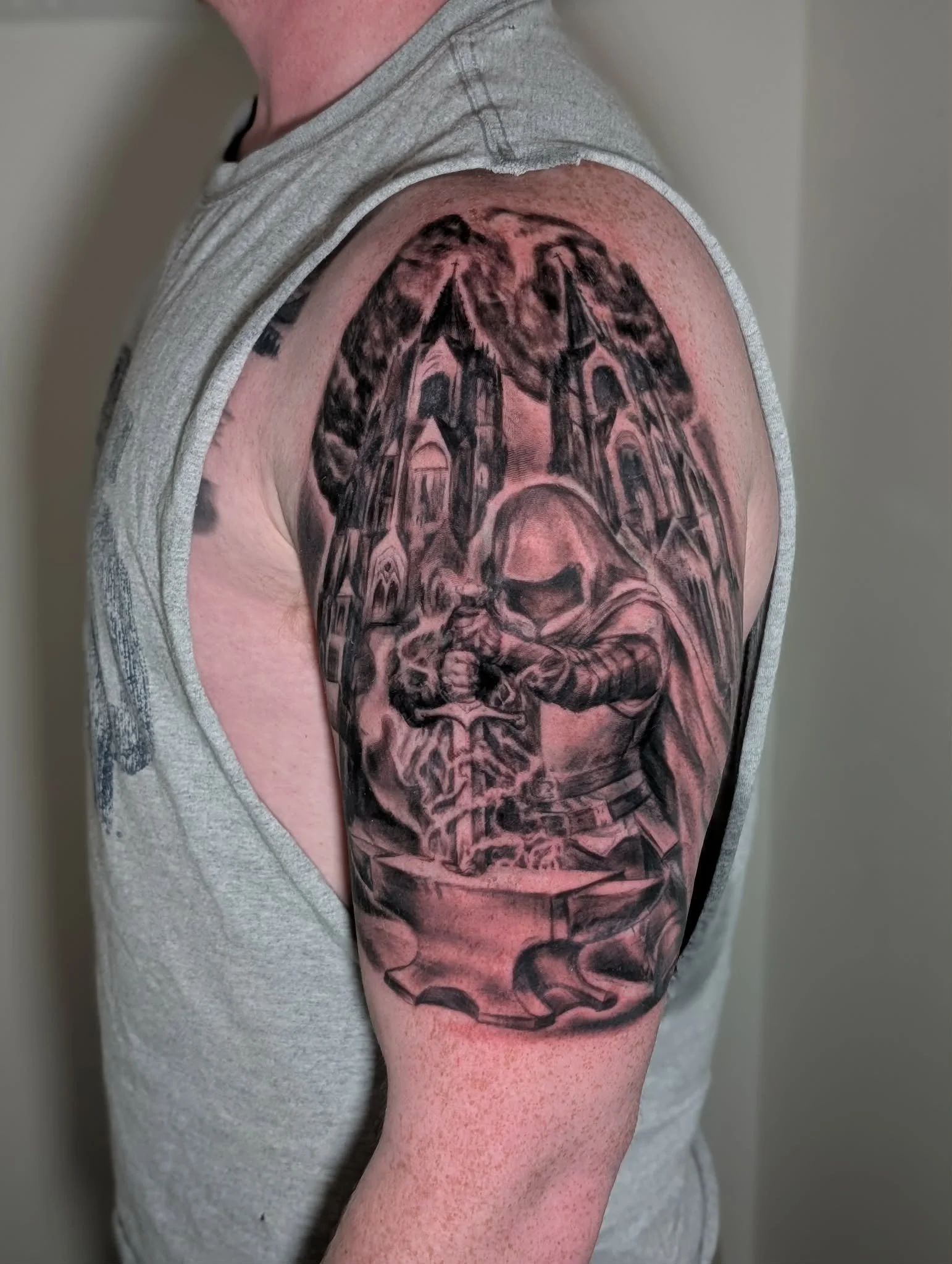 Close-up view of a person's upper arm with a detailed black and gray tattoo depicting a church, trees, and a religious figure.
