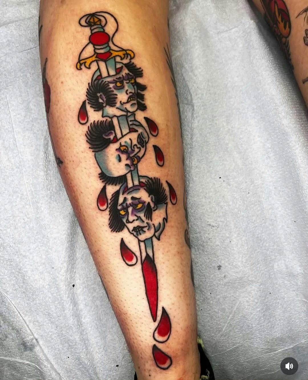 Traditional tattoo of a sword piercing through three faces of a man with long hair and mustaches, with red teardrop-shaped drops beneath each face.
