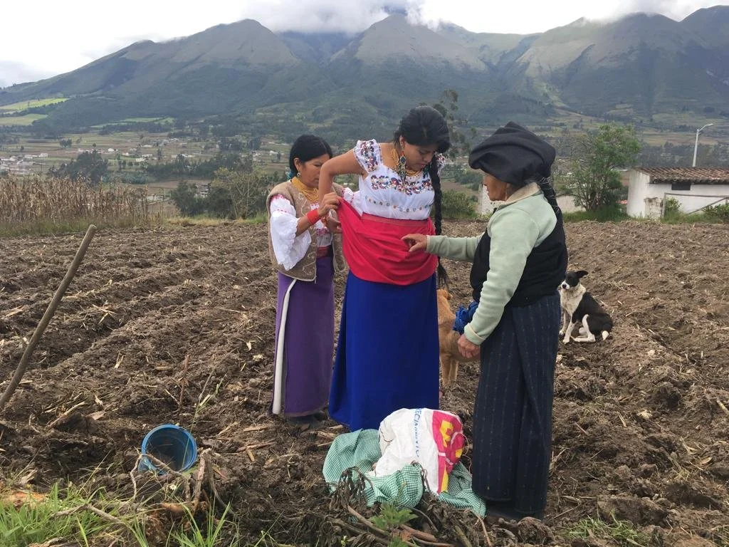 Three women in traditional clothing standing in a field with mountains in the background, one woman is adjusting her dress while another is pointing at her. A black and white dog is sitting on the ground nearby.
