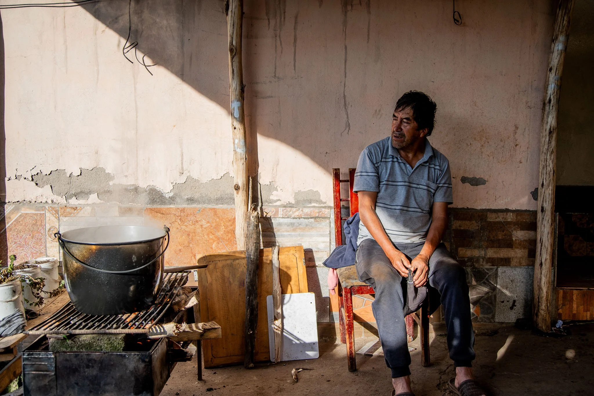 A man sits on a red chair outside a rustic building with a cracked wall, nearby a cooking pot on a makeshift stove, in a rural setting.