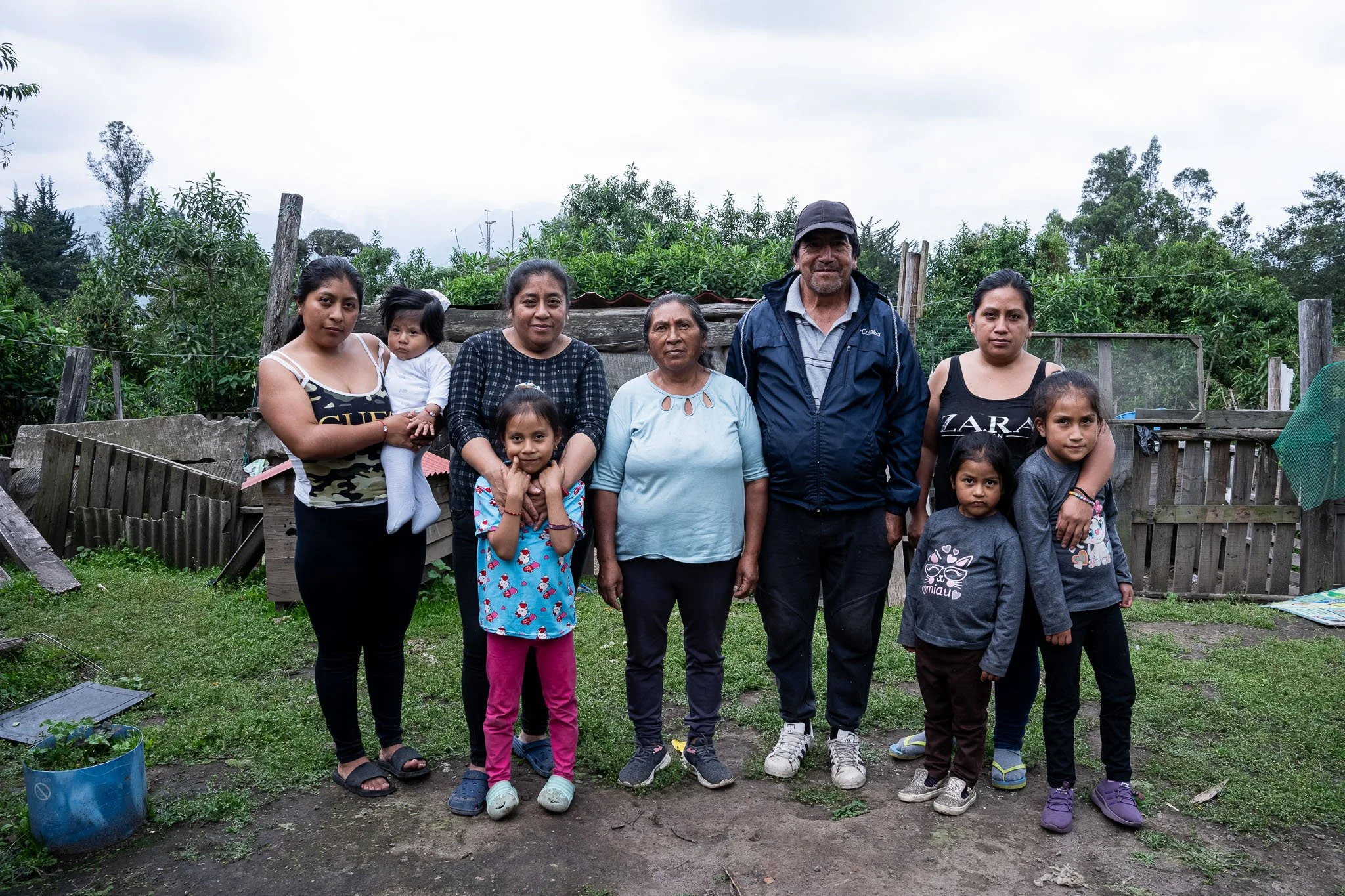 A group of ten people, including adults and children, standing outdoors on a grassy area with a rustic wooden fence and green trees in the background. Chaos with overcast sky.