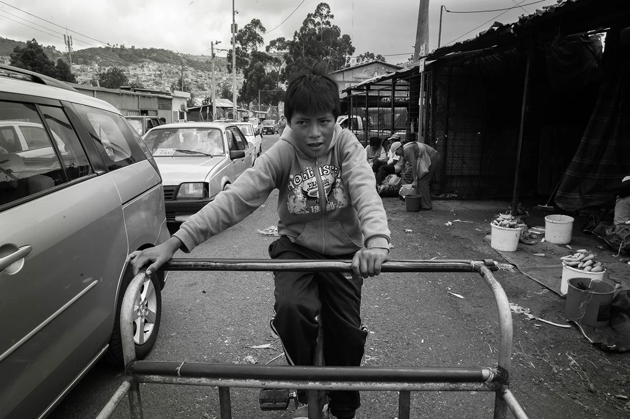 A young boy riding a metal cart on a busy street with cars parked nearby and a market with vendors in the background.