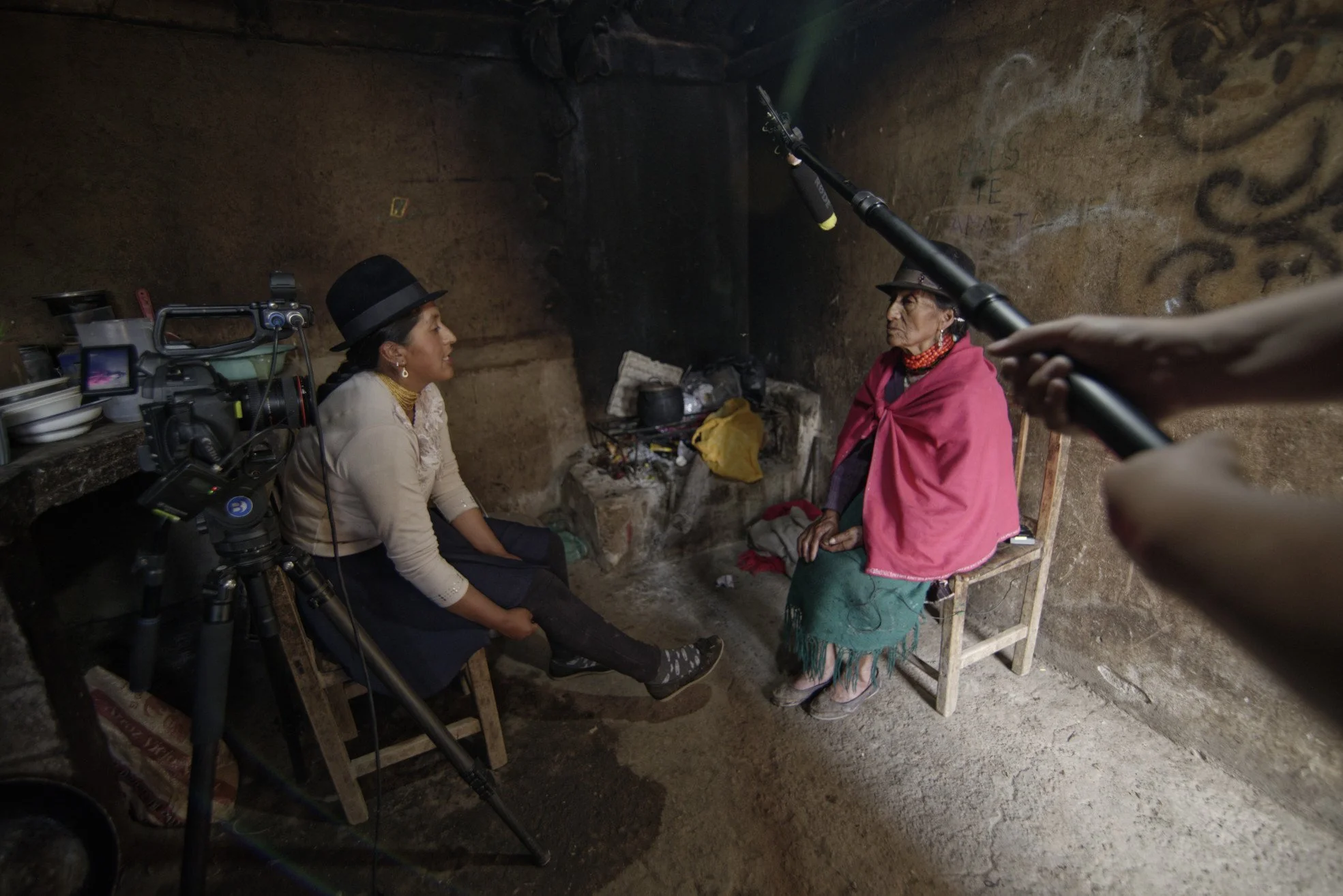 Two people sitting in a rustic room during an interview setup, with one person holding a microphone and a camera on a tripod recording the conversation.