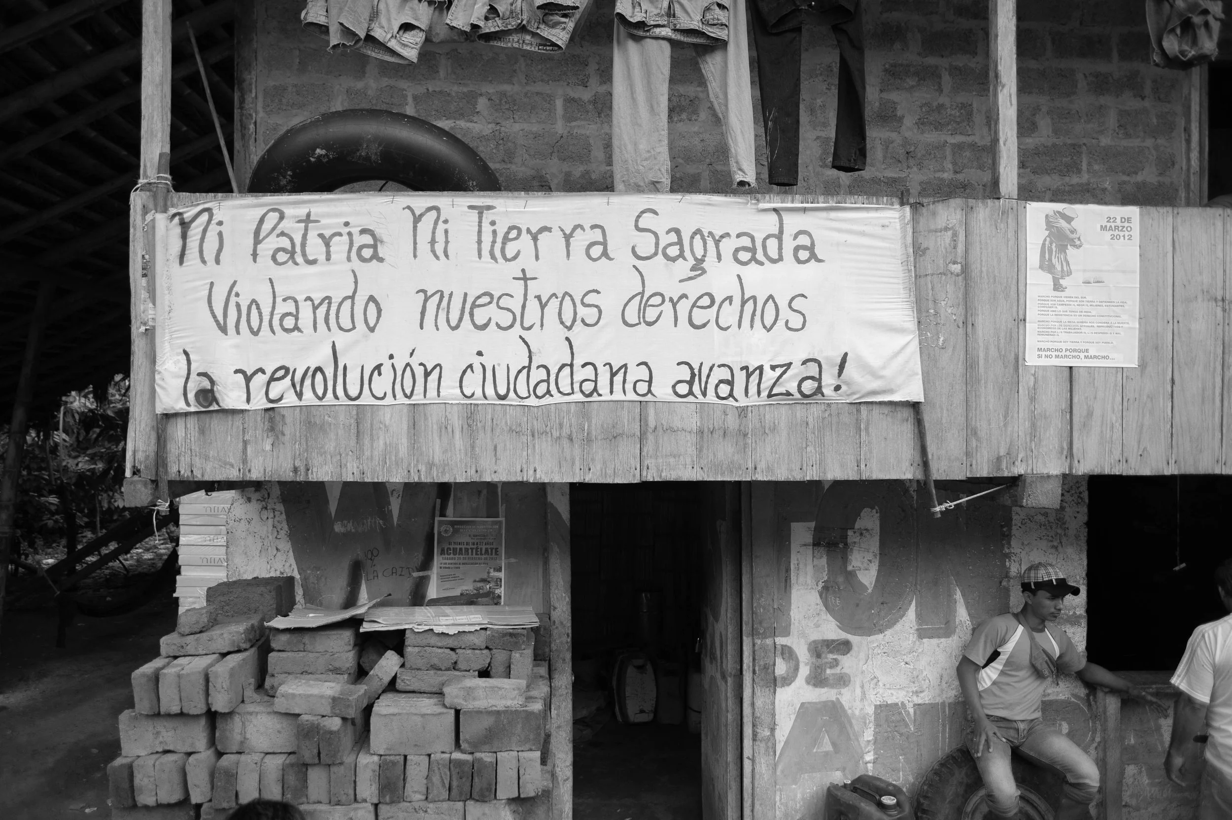 A handmade banner with a message in Spanish hanging on a wooden wall above a garage-like space. The message reads 'Mi patria, mi tierra sagrada violando nuestros derechos la revolución ciudadana avanza!' The scene includes stacked bricks and a man sitting on a tire.