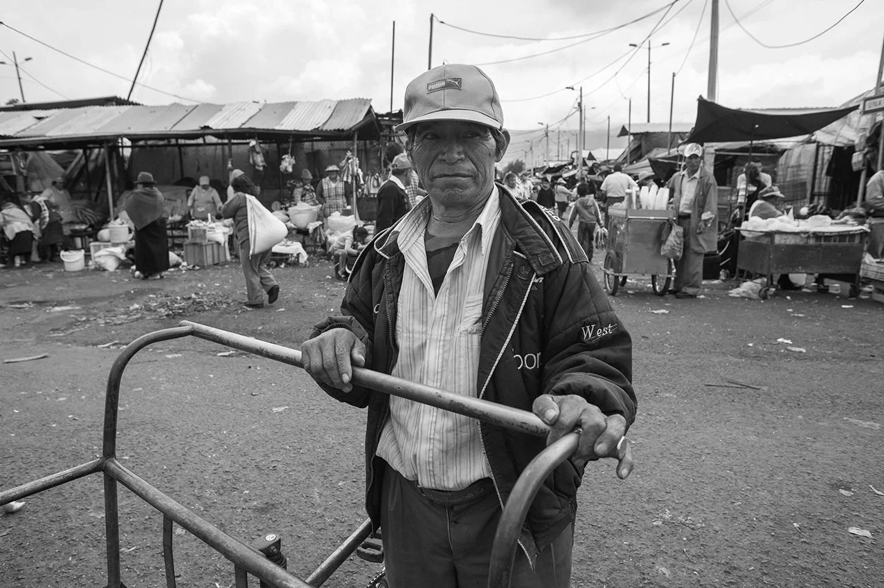 A man standing in a busy outdoor market with a cart, surrounded by many vendors and shoppers, in a black and white photograph.