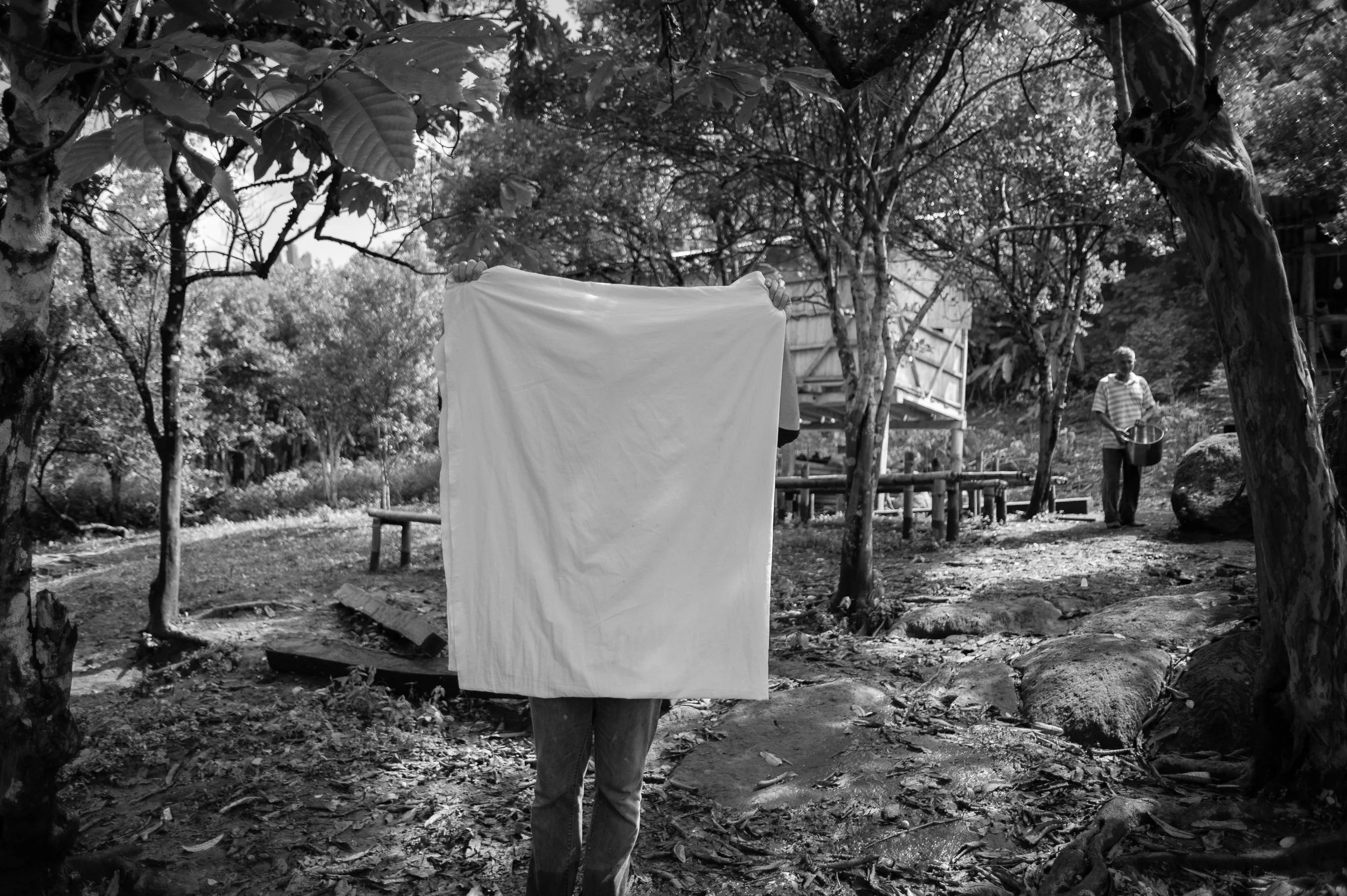 A person holding a large white cloth or sheet in front of their face, standing outdoors in a wooded area, with another person walking in the background holding a pot.