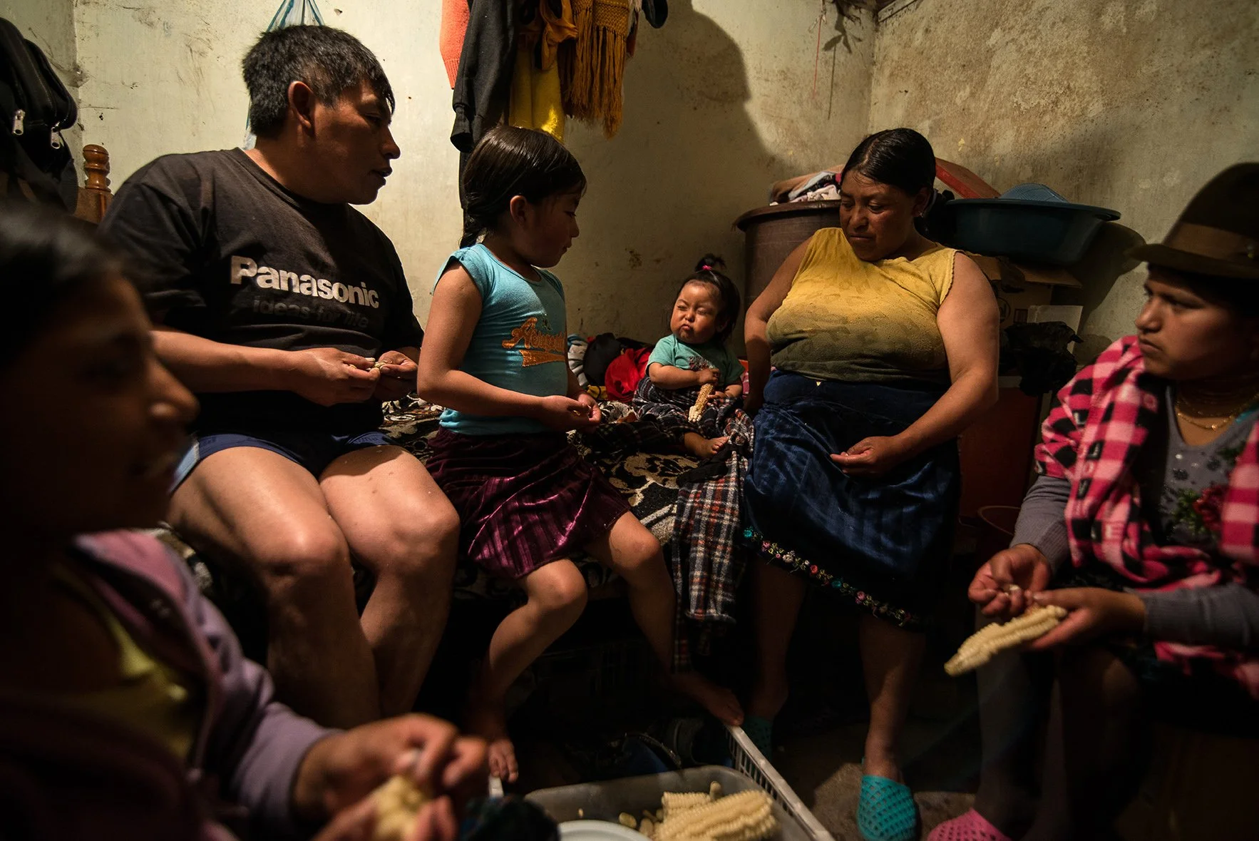 A group of people, including children and women, sitting in a small, dimly lit room with worn walls, some holding bread or corn, engaged in conversation or eating.