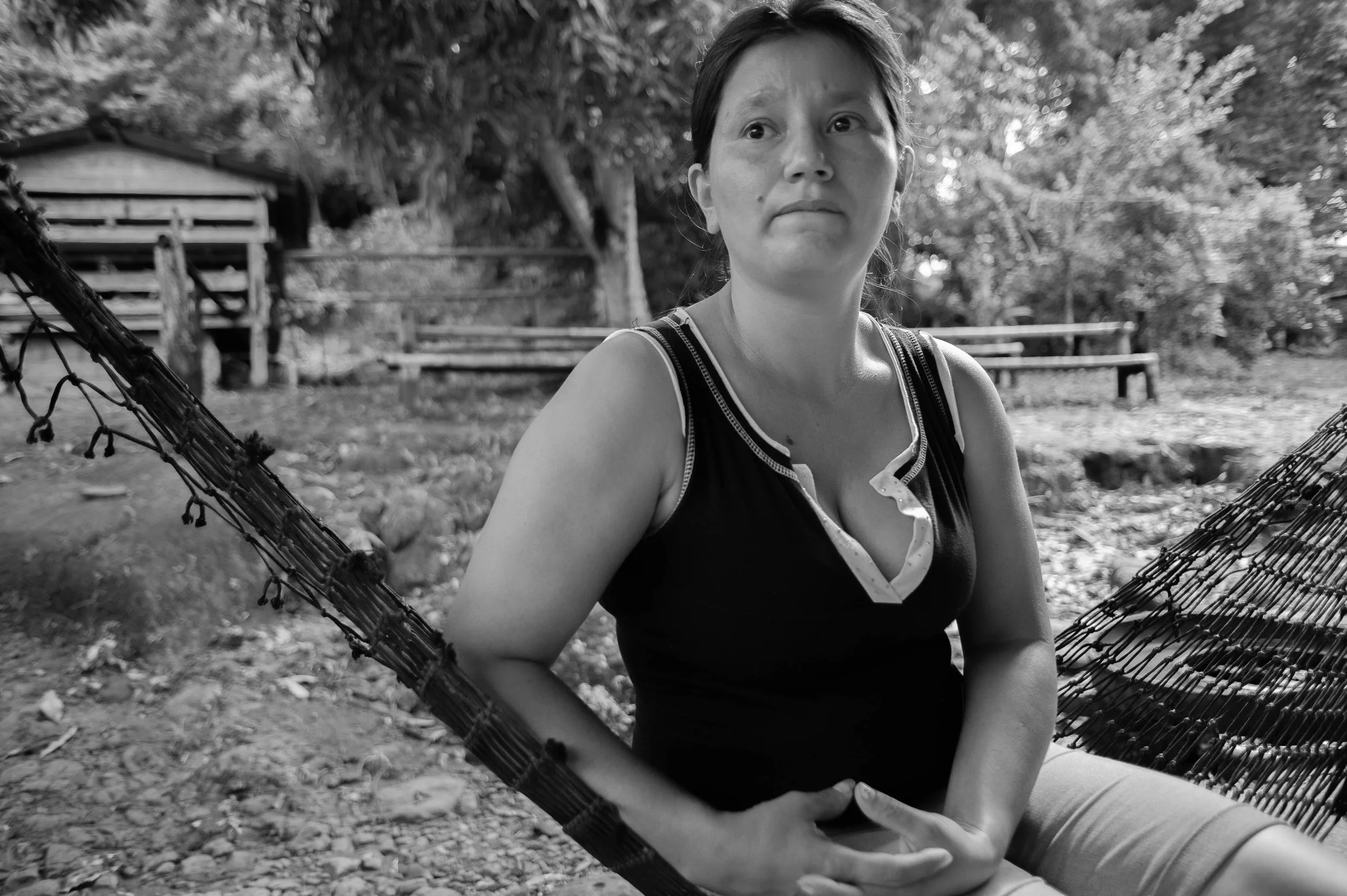 Black and white photo of a woman sitting in a hammock outdoors with trees and wooden benches in the background.