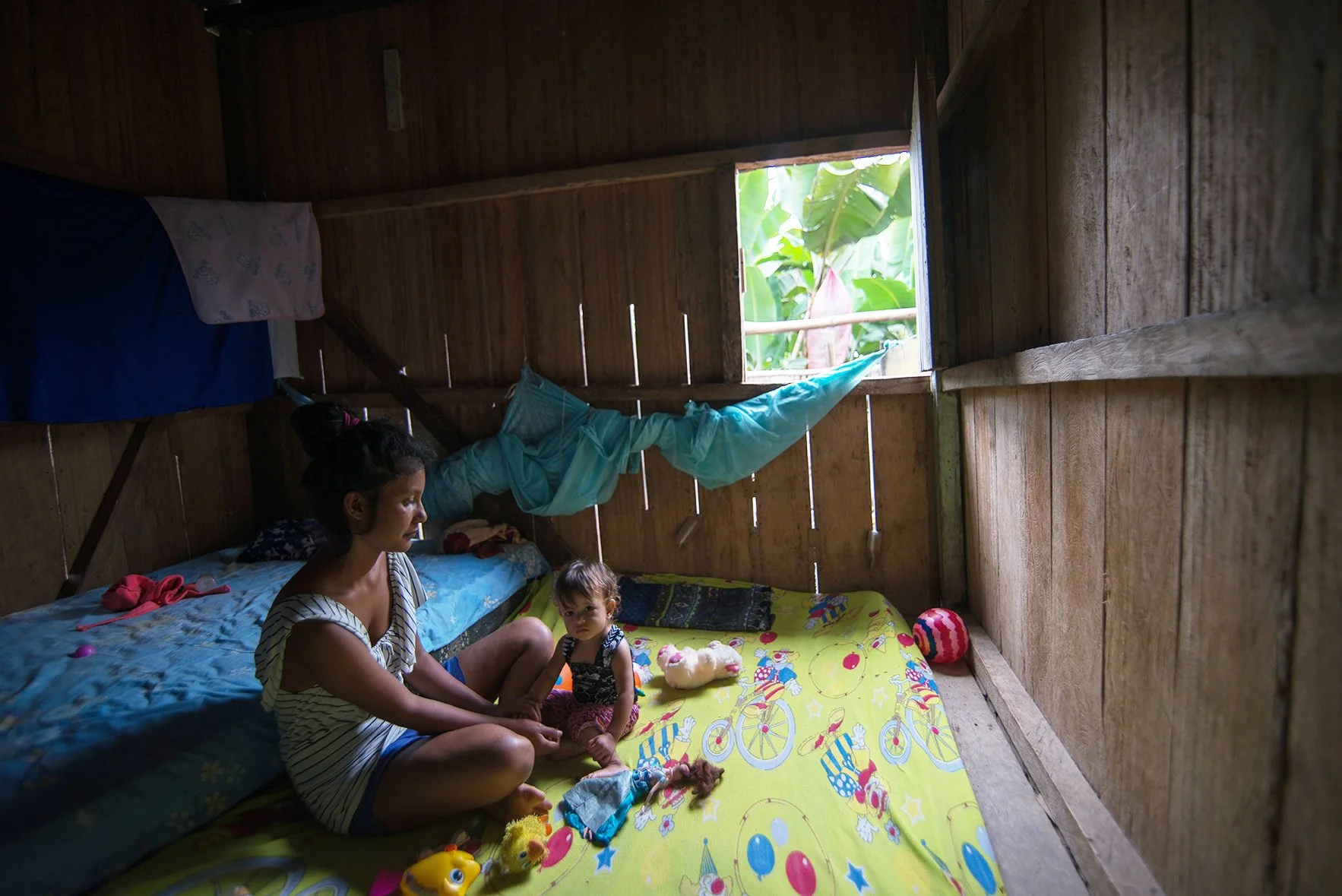 A woman and a young girl sitting on a colorful bed inside a wooden room with a small window showing green foliage outside. The room has toys and stuffed animals on the bed.