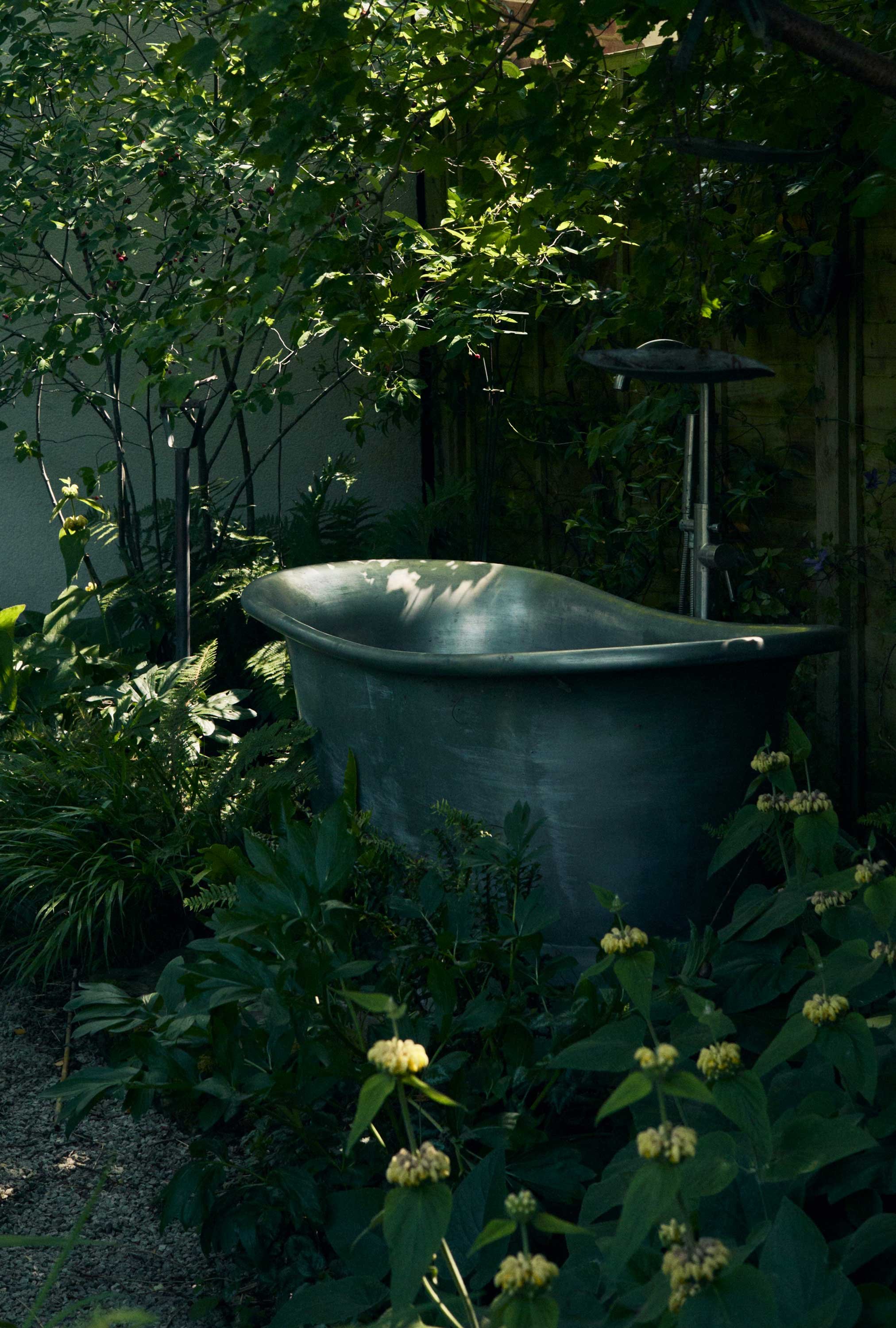 An old bathtub used as a garden planter surrounded by green plants and partially shaded by trees in a backyard garden.