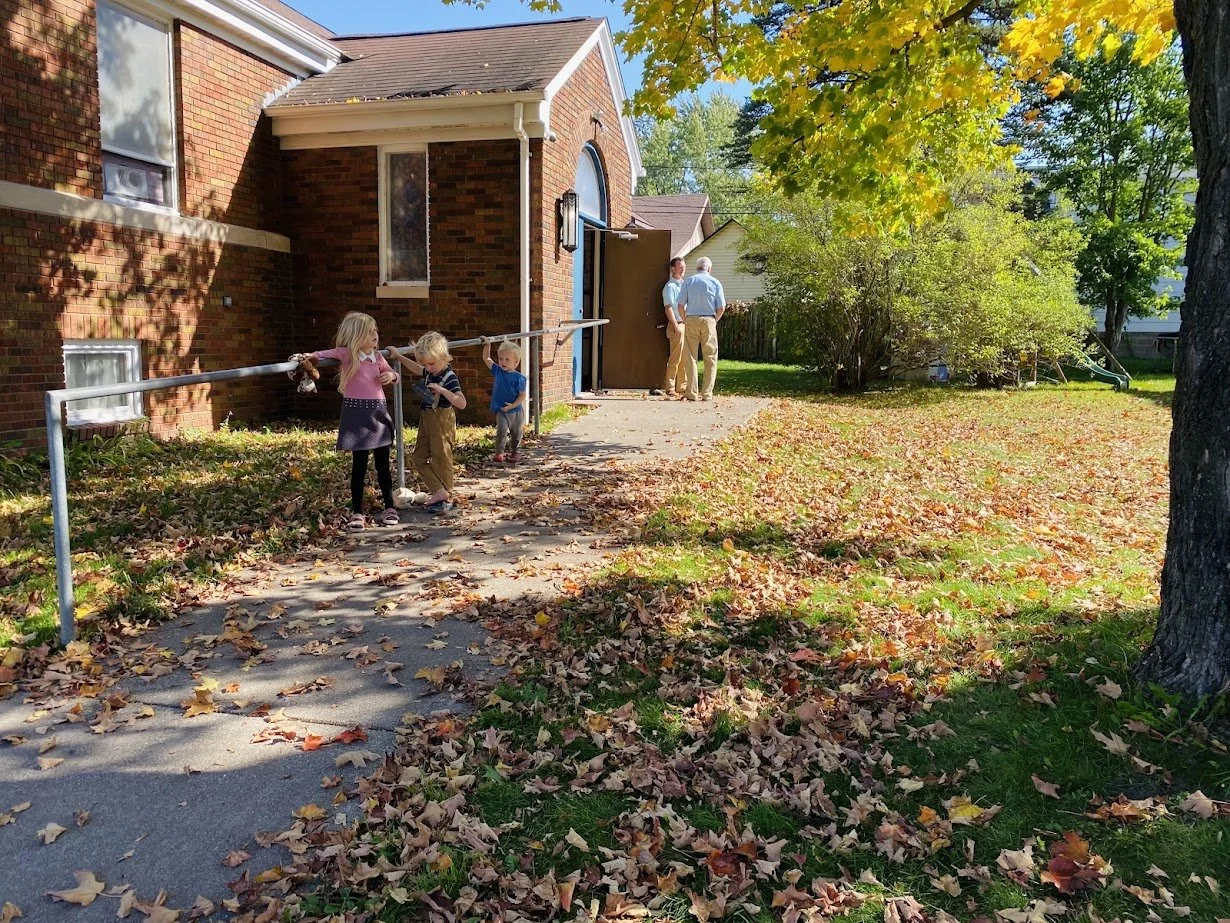 Children picking up fallen leaves outside a brick house on a fall day, with two adults standing near the open house door.